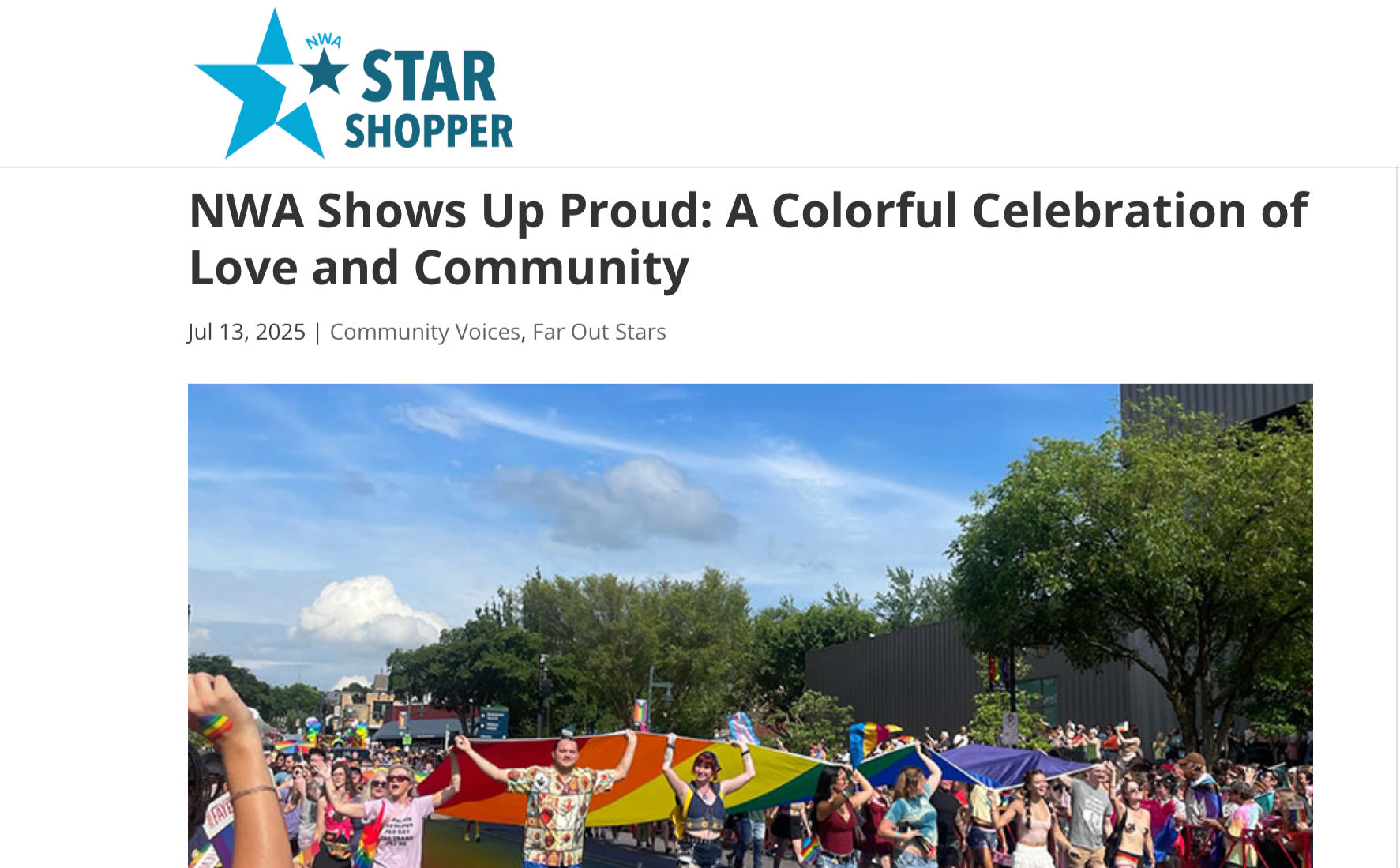 A large crowd of people celebrating at an LGBTQ pride parade, holding a rainbow flag, with some participants wearing rainbow accessories, under a blue sky with clouds, with trees and buildings in the background.