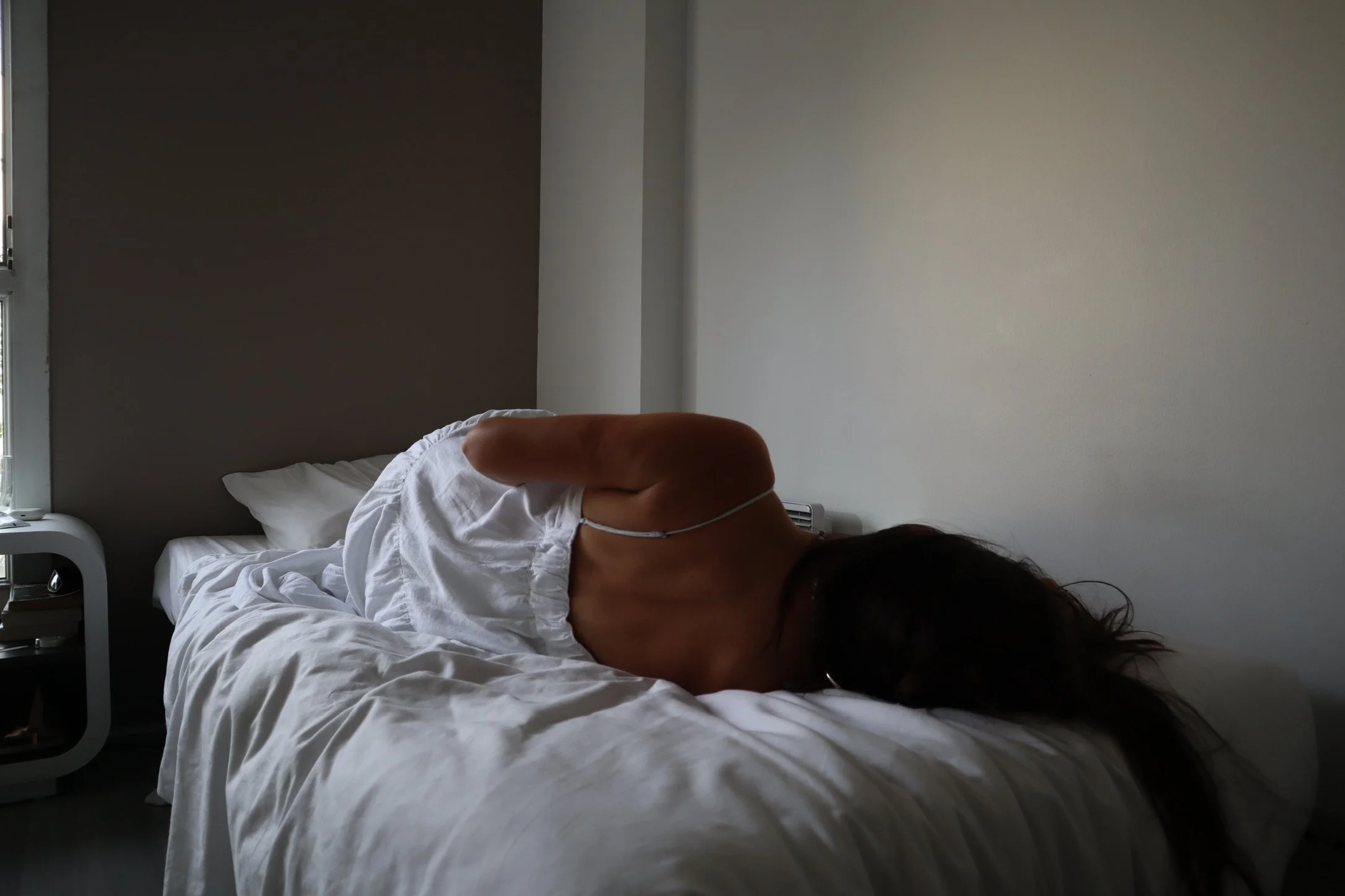 A woman lying on her side on a bed in a minimalistic bedroom, with dark hair partially covering her face, wearing a white sleeveless top.