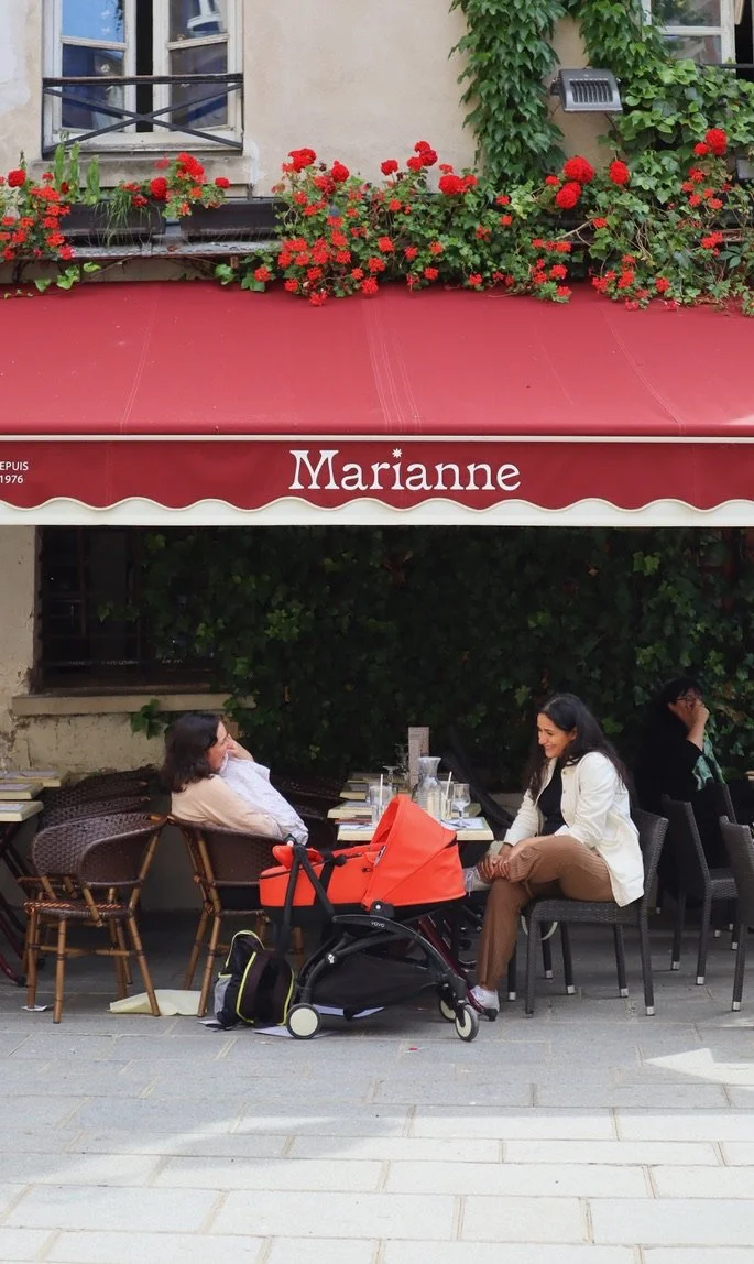 Women sitting at an outdoor cafe table with a red stroller and backpack, under a red awning with the name "Marianne," flowering plants above, and a building with windows behind.
