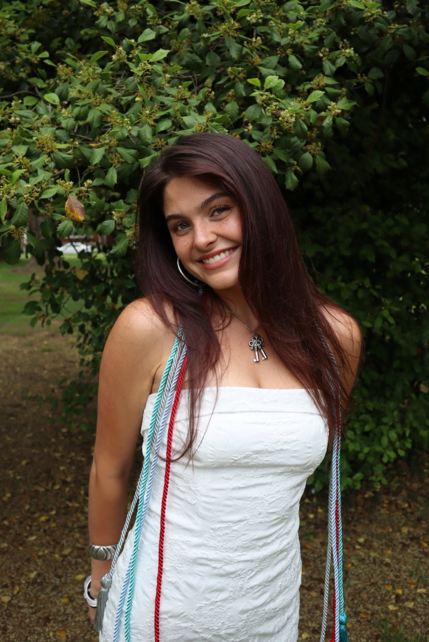 A young woman with long dark hair, smiling, wearing a white dress and graduation cords, standing outdoors in front of green foliage.