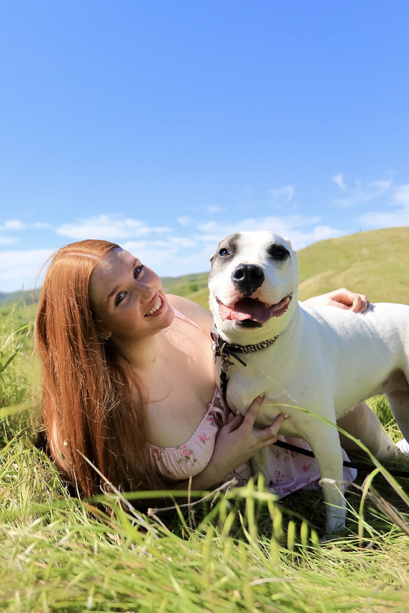 A woman with long red hair lying on green grass in a field, smiling, with a white dog that has black markings on its face, also smiling, under a blue sky with some clouds.