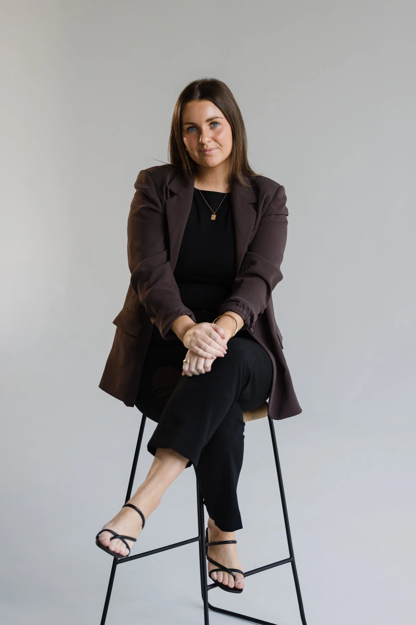 A woman with shoulder-length brown hair sitting on a tall stool against a plain light-colored background. She is wearing a dark blazer, black top, black pants, and black strappy sandals, with a gold necklace and rings visible.