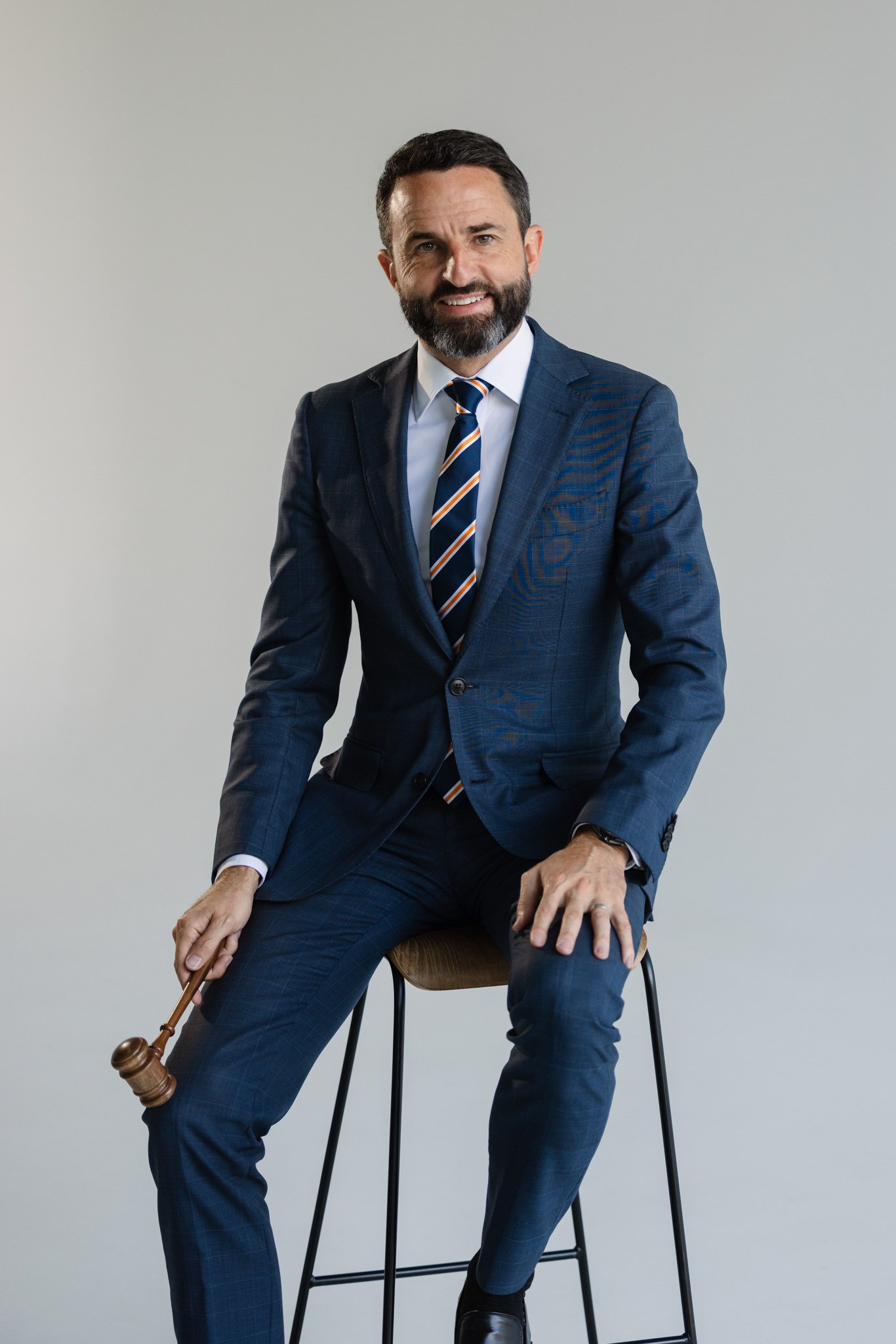 Man in a blue checkered suit, sitting on a stool, holding a gavel, with a neutral background.