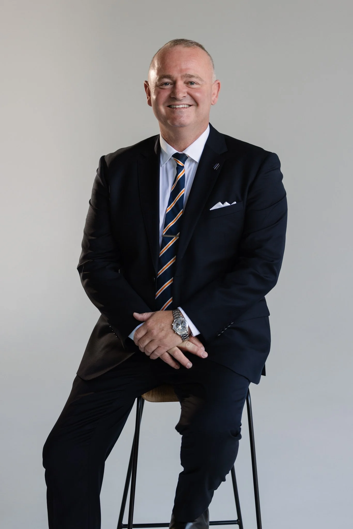 A middle-aged man in a dark suit, white shirt, and striped tie sitting on a stool against a plain gray background.