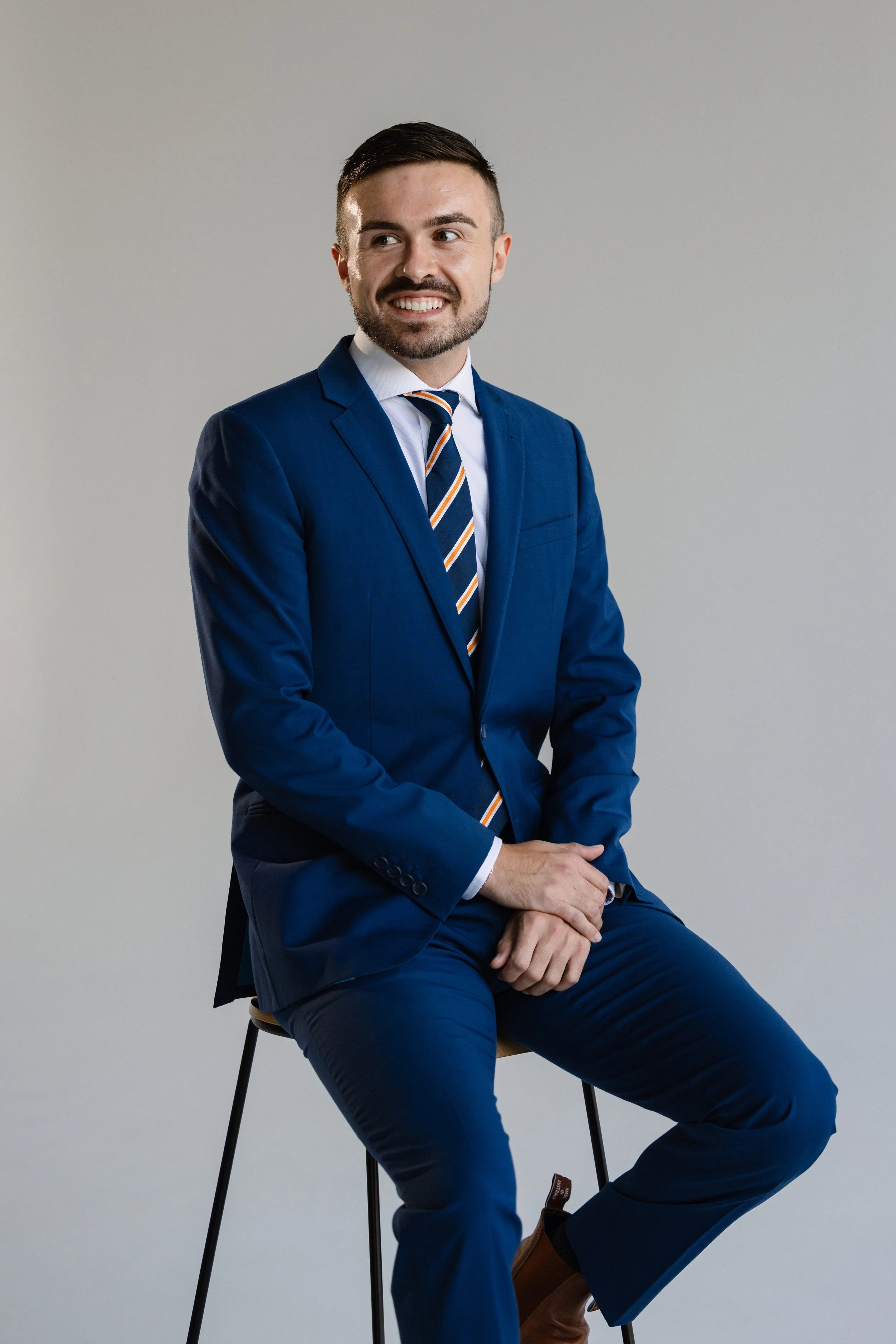 A man in a blue suit, white shirt, and striped tie sitting on a stool with hands clasped, smiling, against a plain gray background.