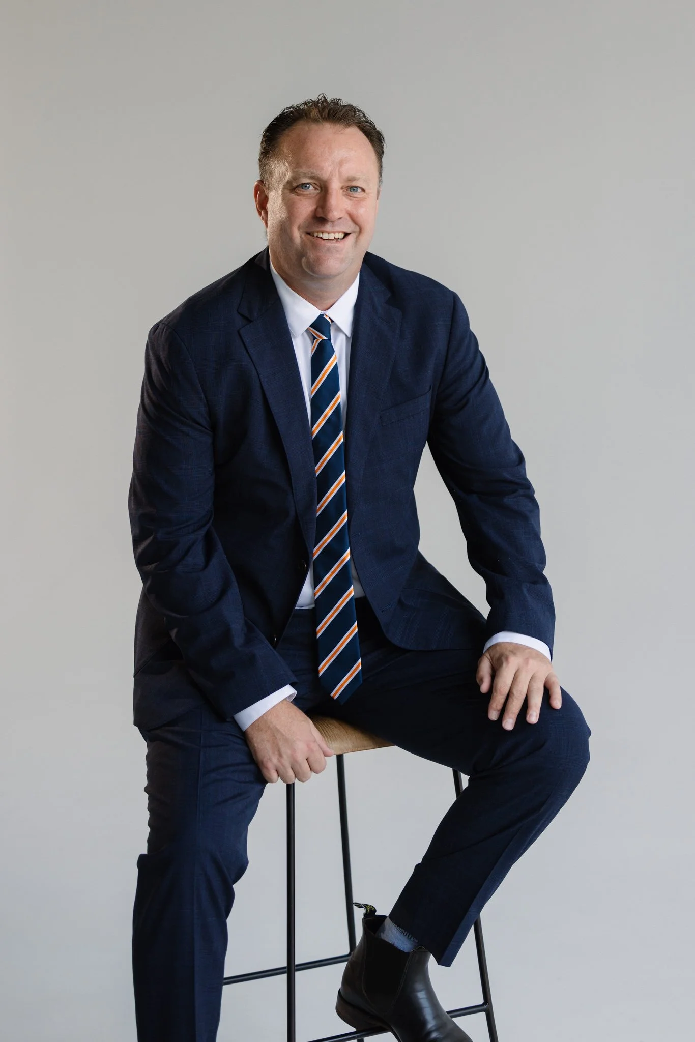 A man in a dark blue suit and tie sitting on a stool against a plain light gray background.