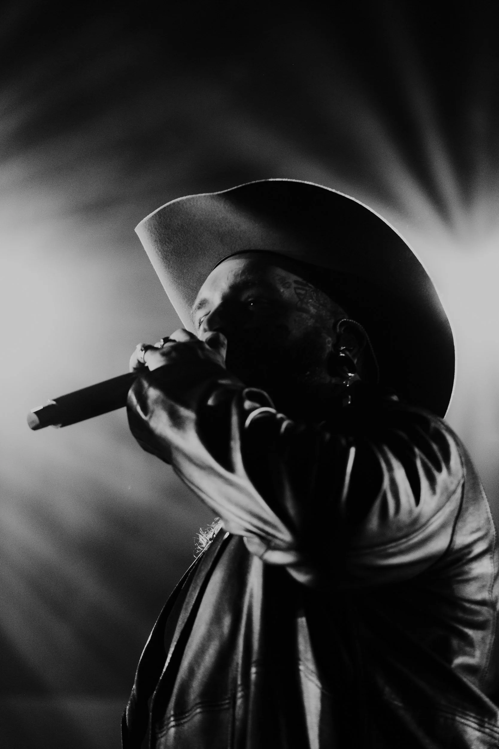 Black and white photo of a male singer wearing a cowboy hat and leather jacket, holding a microphone close to his mouth, performing on stage.