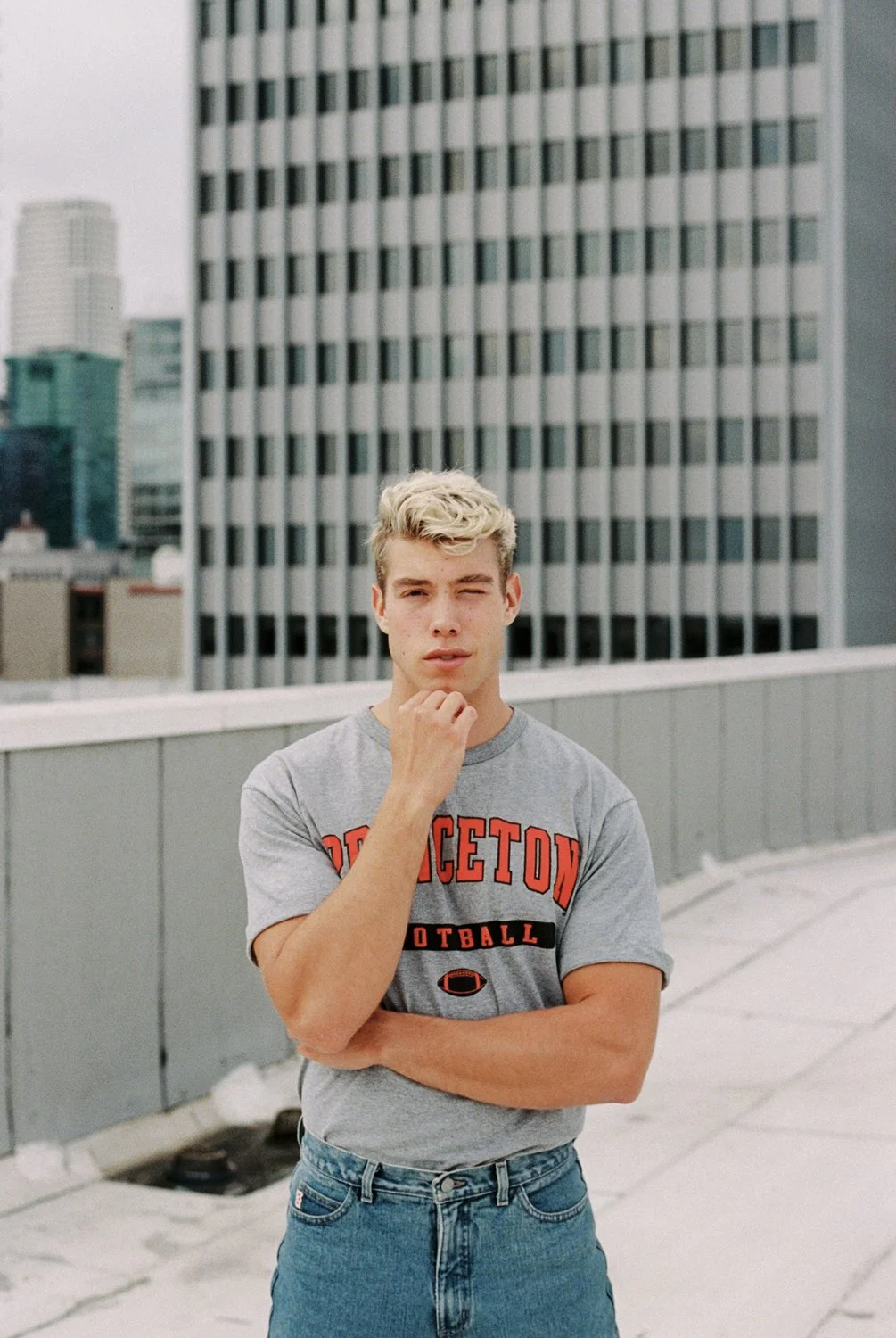 A young man with blonde curly hair standing on a rooftop in an urban area, wearing a gray Princeton football T-shirt and blue jeans, with city skyscrapers in the background.