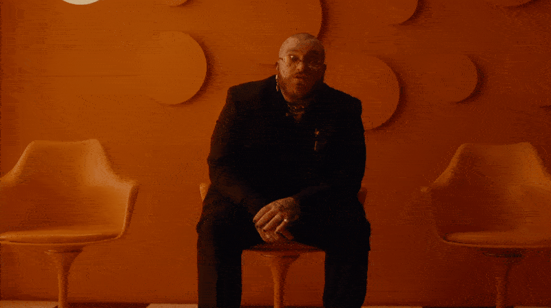 A man in black sitting on a stool in a dimly lit room with a modern, circular wooden wall design, flanked by two similar chairs.