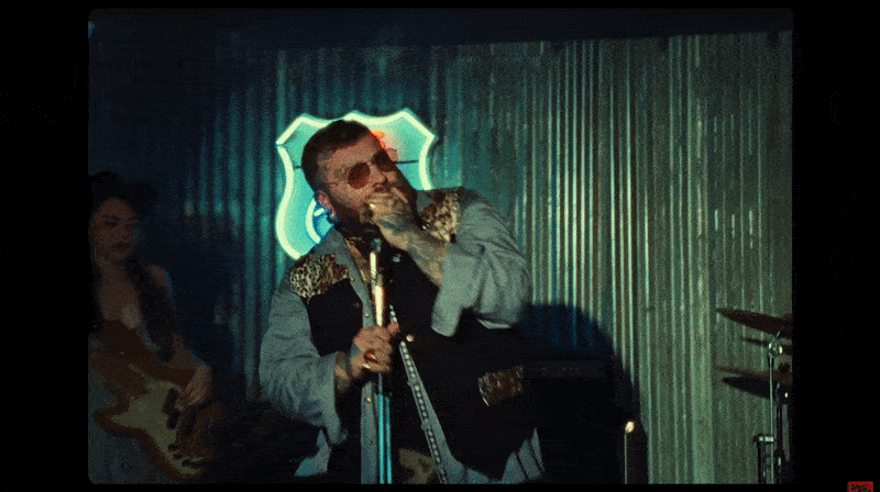 A man with sunglasses singing and playing a microphone on a stage with a metal wall background, illuminated by a neon sign behind him.