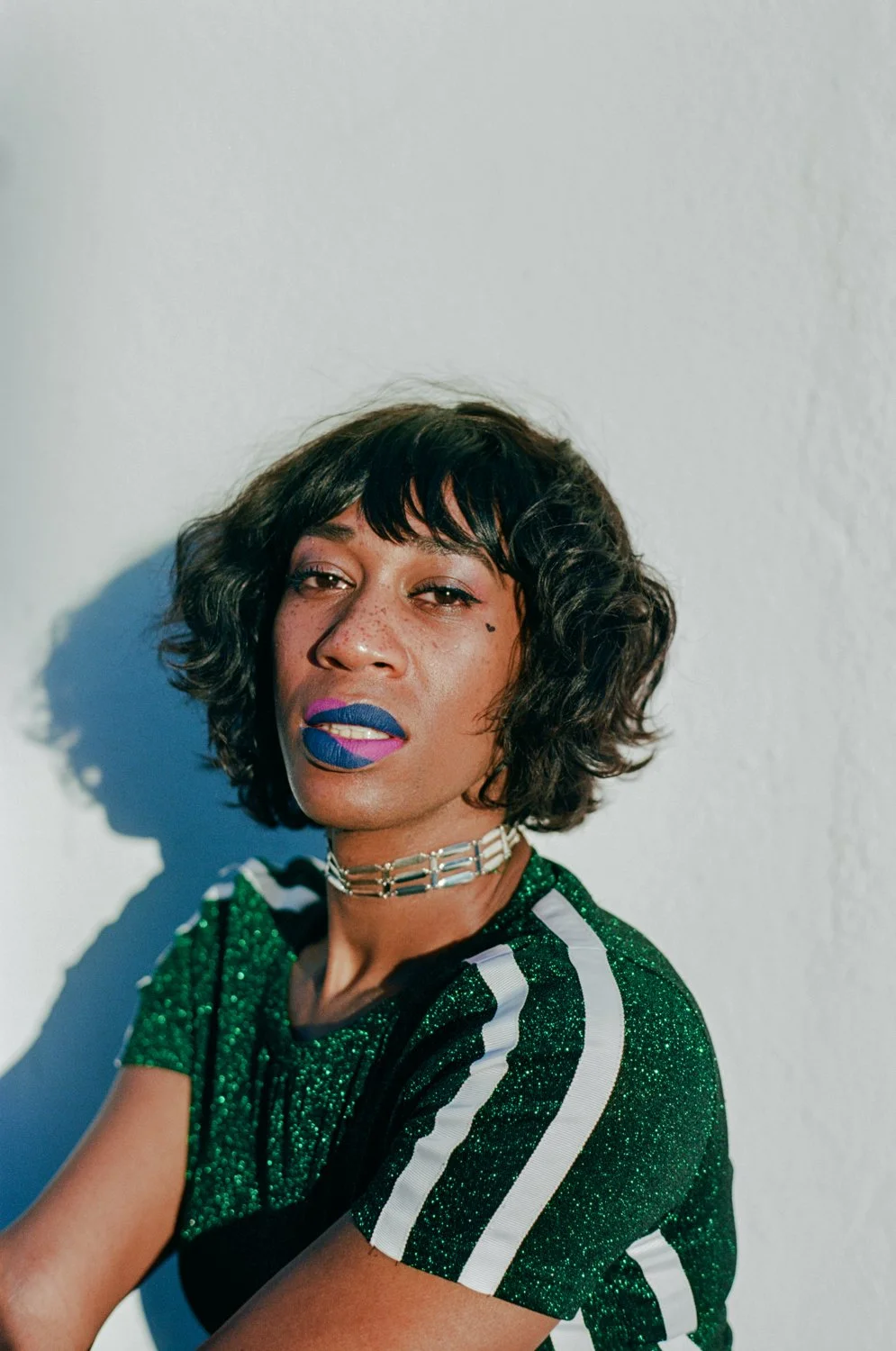 A woman with dark curly hair and colorful makeup, wearing a green glittery top with white stripes, a silver choker, and bold lipstick, standing against a white wall.