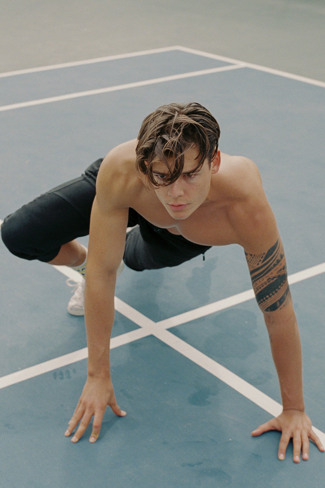 A shirtless young man with tattoos on his right arm, wearing black shorts and white sneakers, in a push-up position on a blue indoor sports court with white lines.