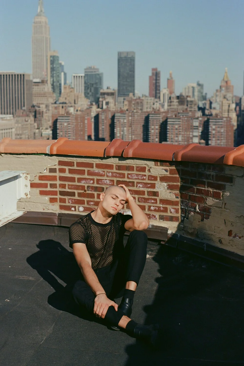 A young person with a buzz cut sitting on a rooftop with a city skyline in the background. They are wearing a black mesh shirt, black pants, and black shoes, with one hand in their hair and eyes closed.