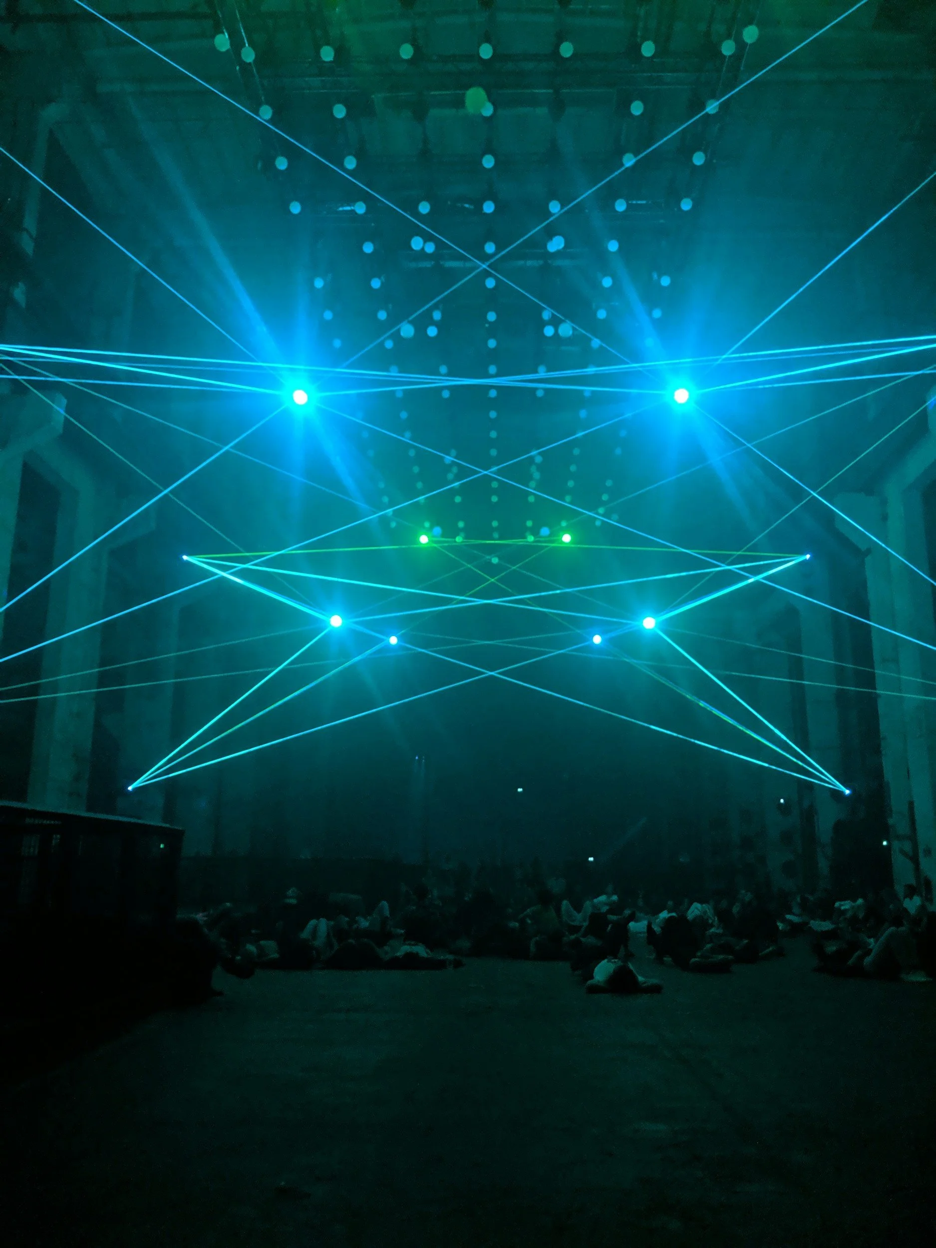 Dark indoor space with colorful laser lights projected overhead, creating geometric patterns. People are seated or lying on the floor in the foreground.