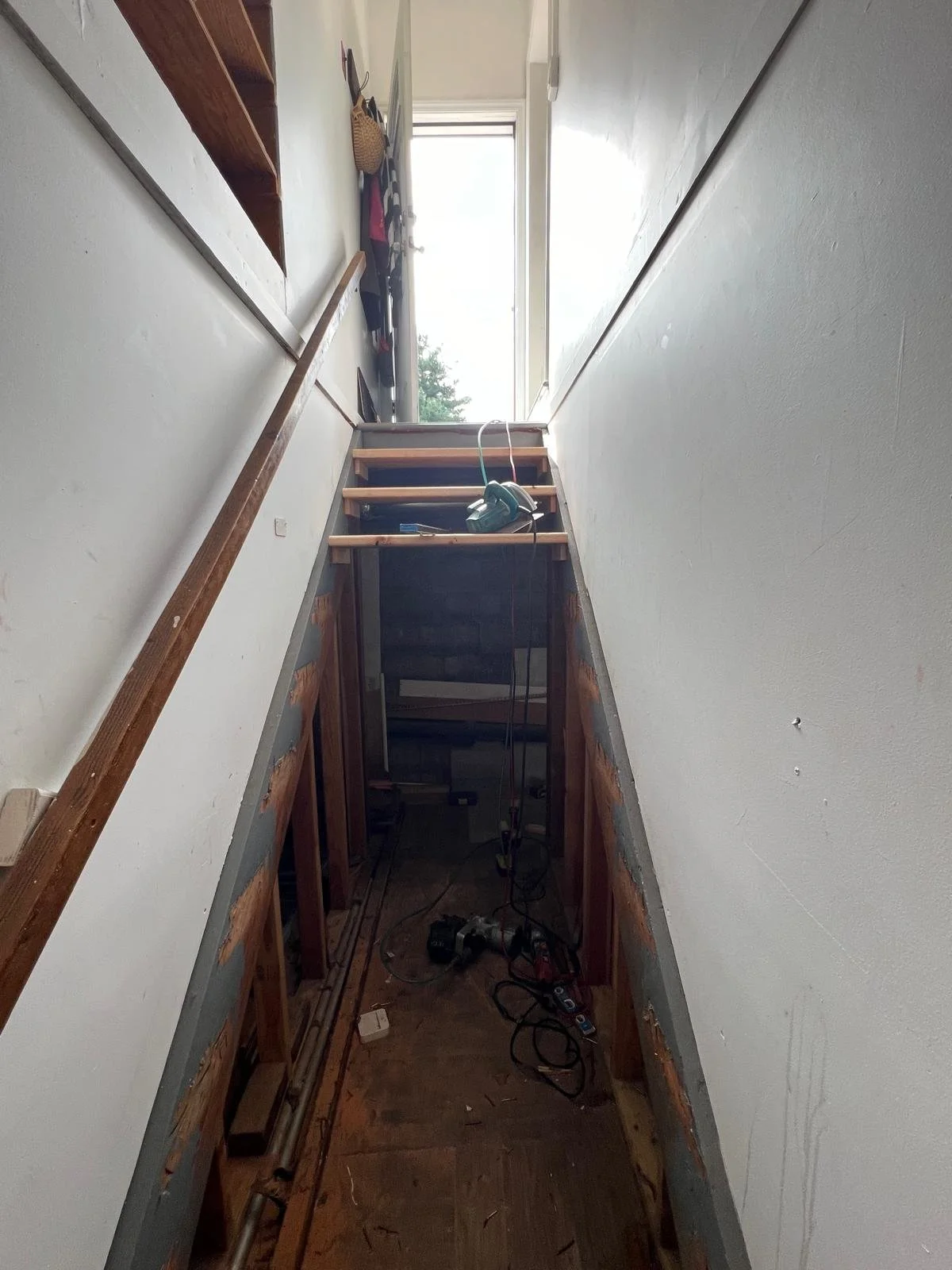 View of a narrow hallway with stairs being built, showing exposed wooden framing, tools, and construction materials, leading to an open door with sunlight coming in.