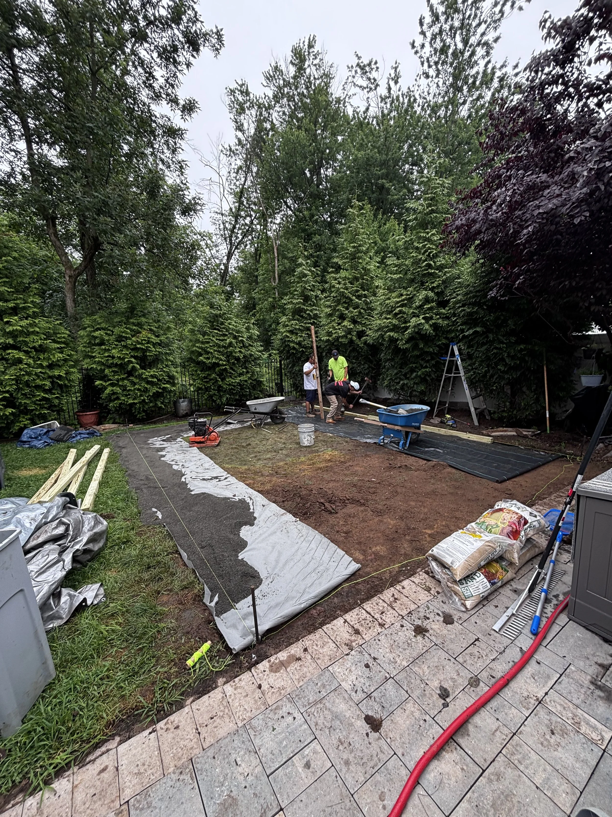 People working on a backyard landscaping project, laying down a gravel or stone pathway, with tools and materials scattered around, surrounded by trees and shrubs.