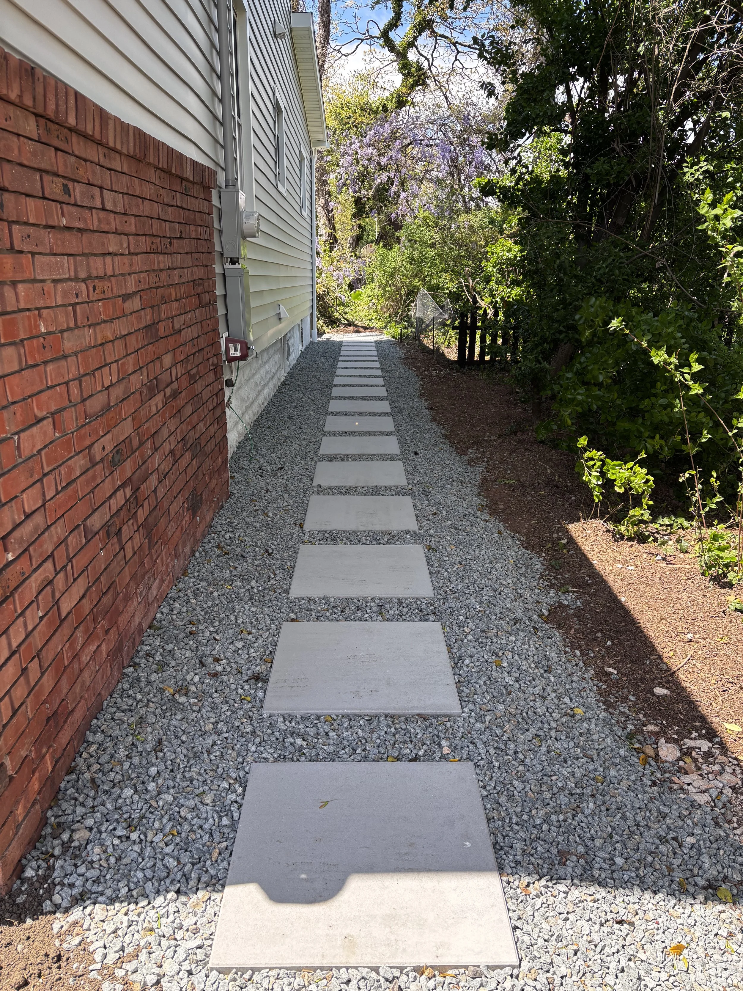 Narrow outdoor walkway beside a house with a brick and siding exterior, featuring concrete stepping stones on gravel, bordered by green plants and trees.