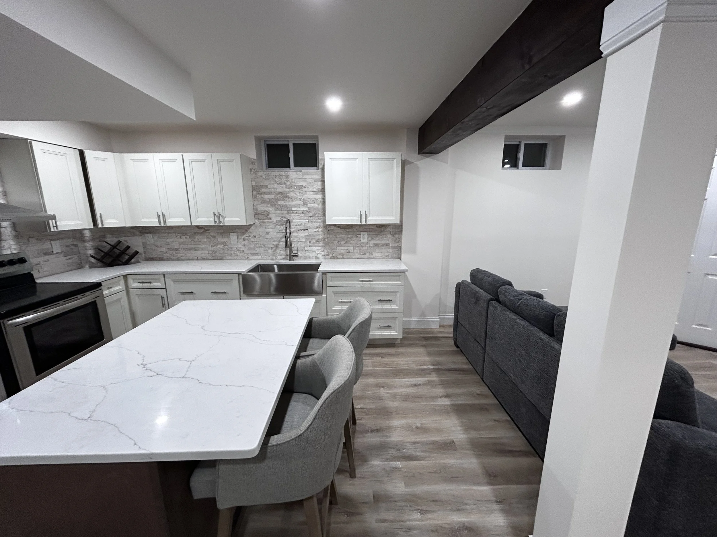 A modern kitchen with white cabinets, a marble countertop island, stainless steel sink, black stove, and gray upholstered chairs. The kitchen has a backsplash of light gray brick and wooden flooring. There is a gray sofa in the adjacent living area w