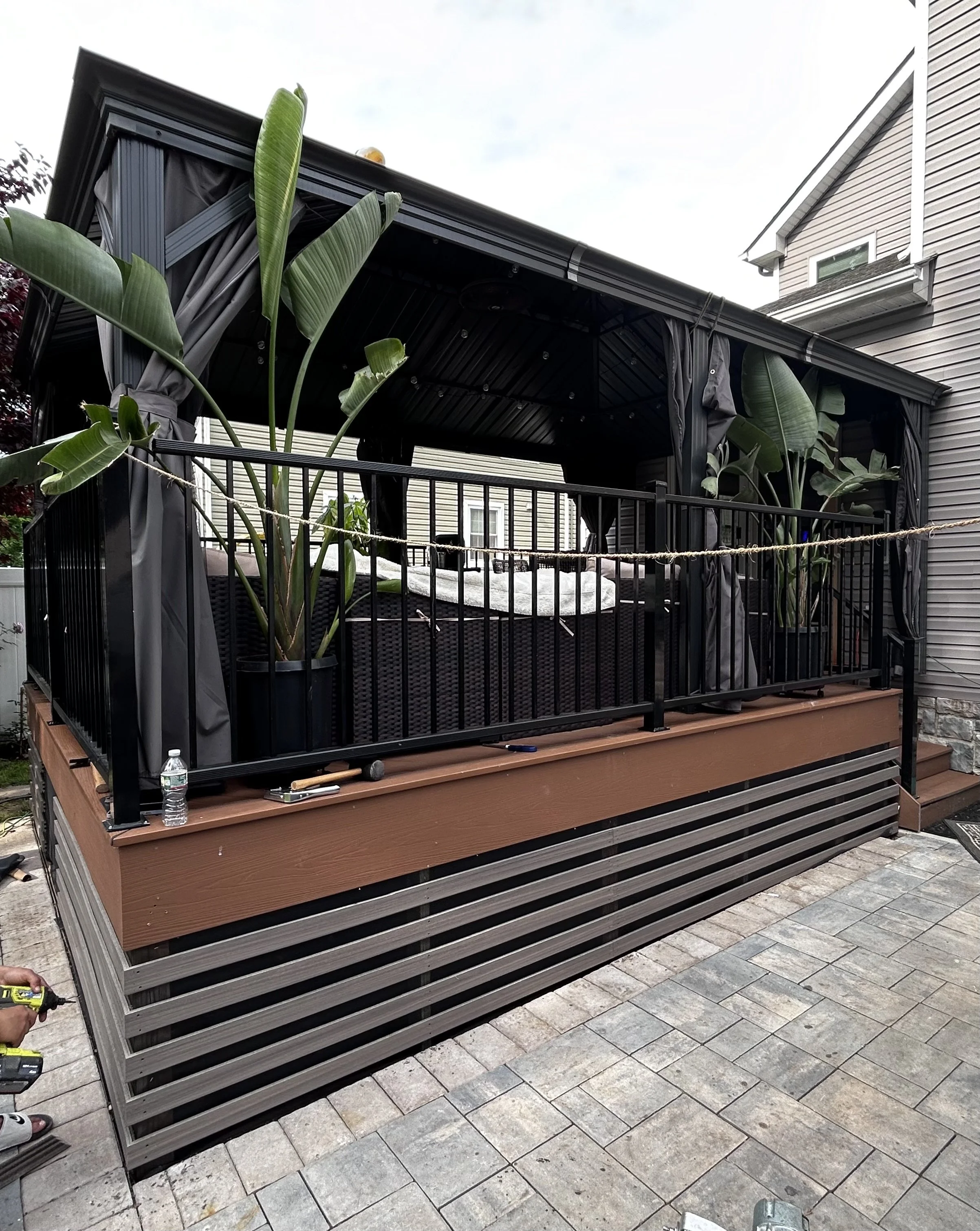 A backyard patio with a raised deck and a screened gazebo. The gazebo has black framing, black curtains tied back, and large green plants inside. The patio has gray paver stones.