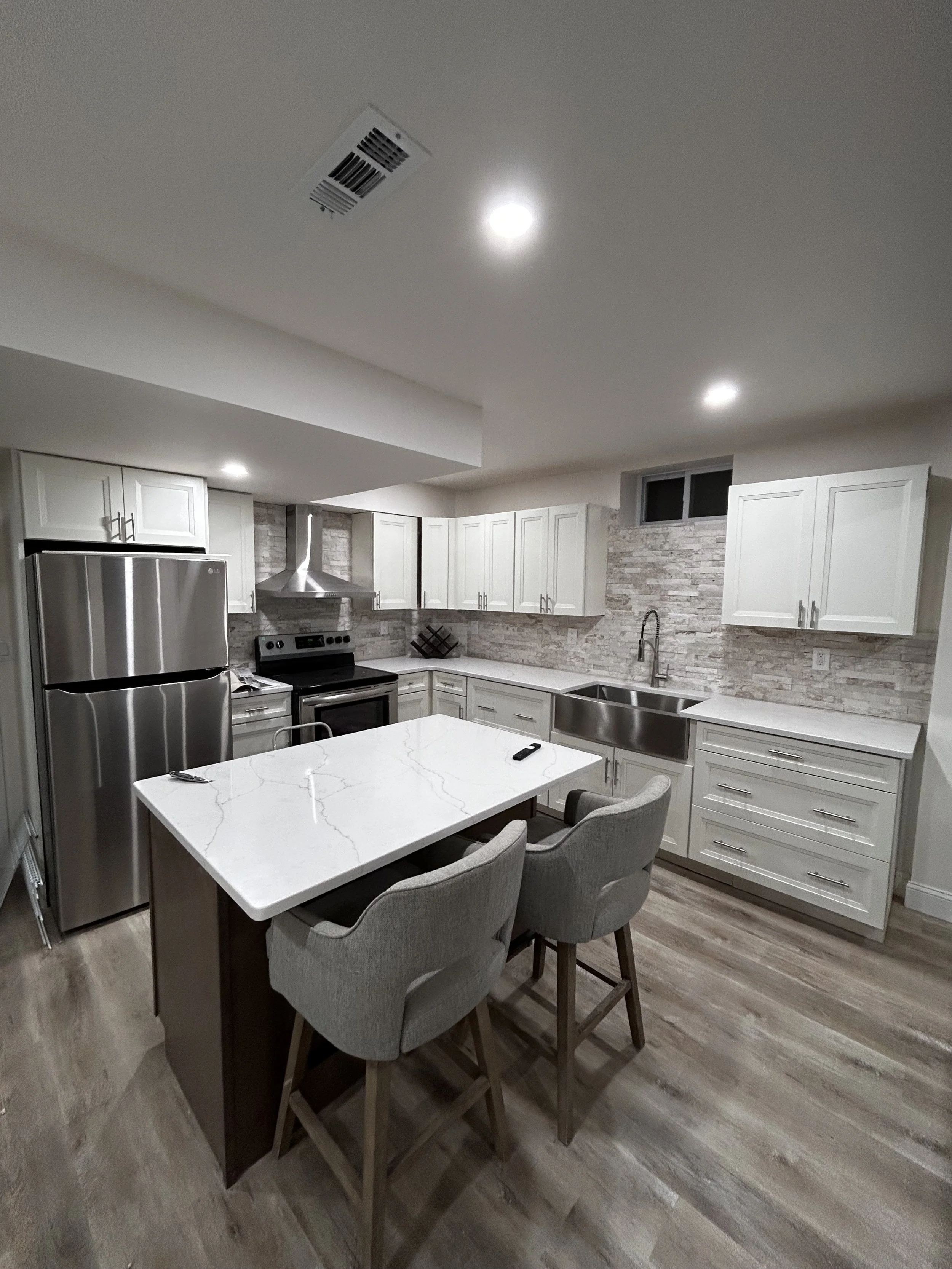 Modern kitchen with white cabinets, stainless steel appliances, a marble island, and light wood flooring.