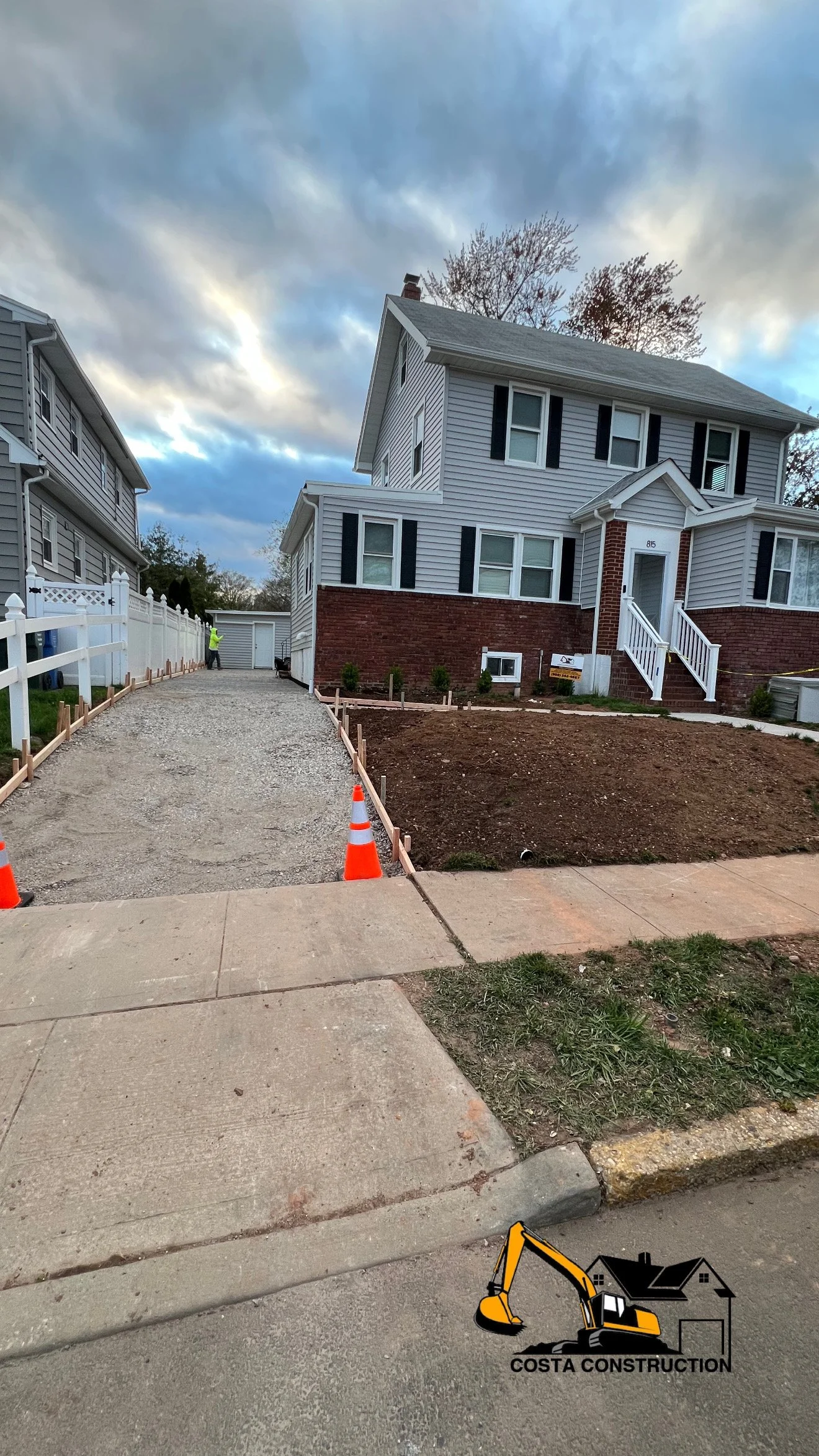 Residential construction site with a gravel driveway, sidewalk, and a house with a brick foundation and white siding. Construction cones and fencing indicate ongoing work.