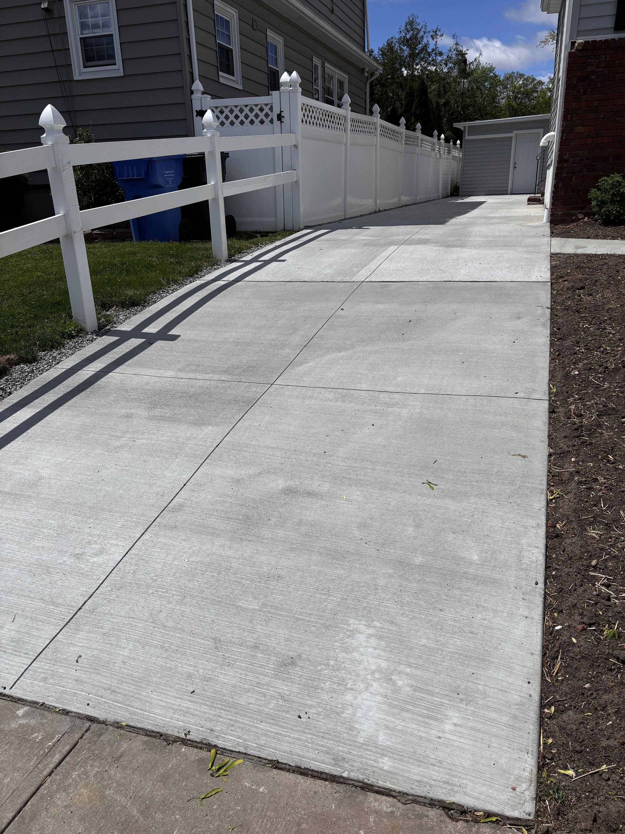 Clean concrete driveway with a slight incline, bordered by a white fence on the left and garden beds on the right, leading to a garage in the background with a partly cloudy sky overhead.