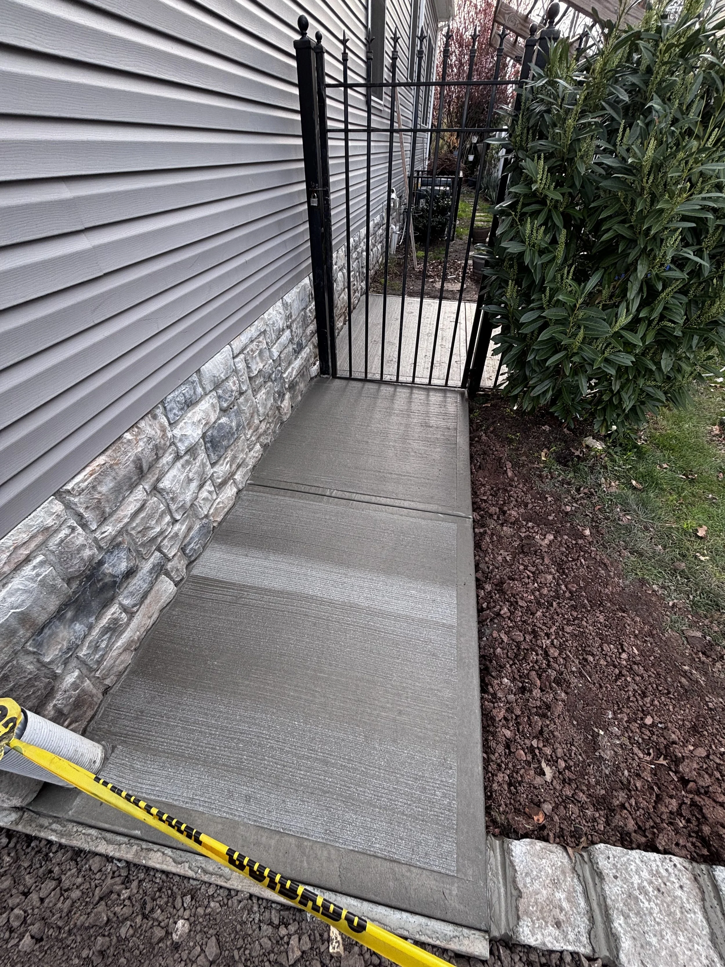Newly poured concrete sidewalk in front of a house, bordered by a stone edge, with a black metal gate and shrubbery on the right.