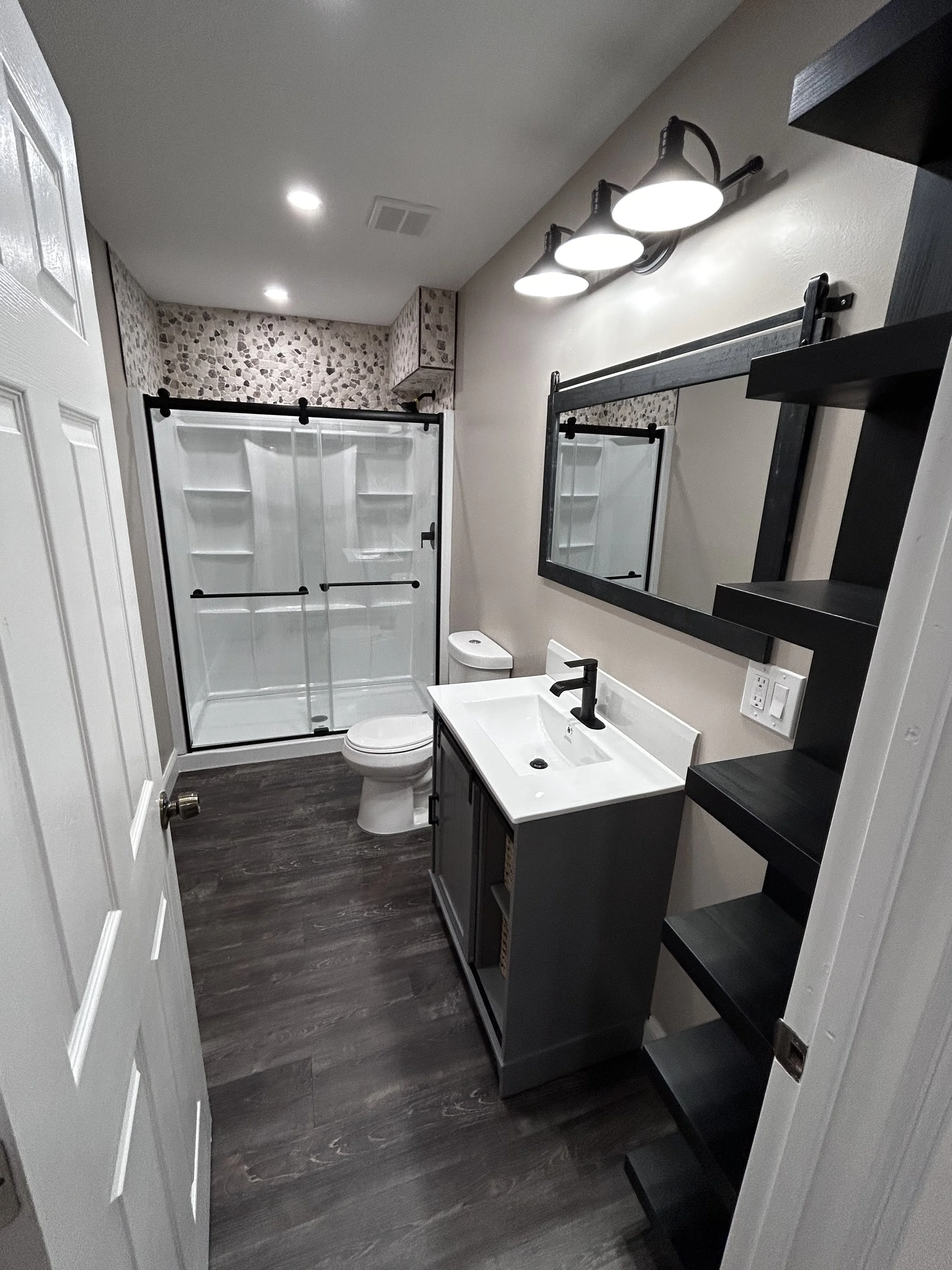 Modern bathroom with a shower, toilet, dark vanity with a sink, mirror, black light fixture, open black shelving, and dark wood flooring.