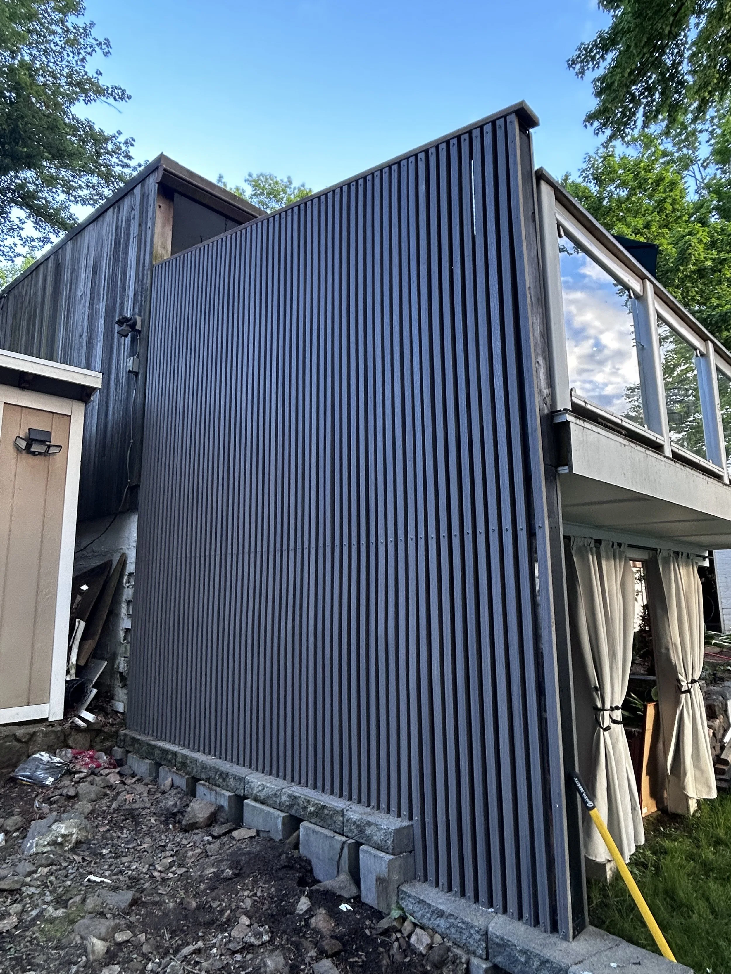 Modern house with vertical dark siding, large windows, and a balcony with curtains, surrounded by trees under a partly cloudy sky.