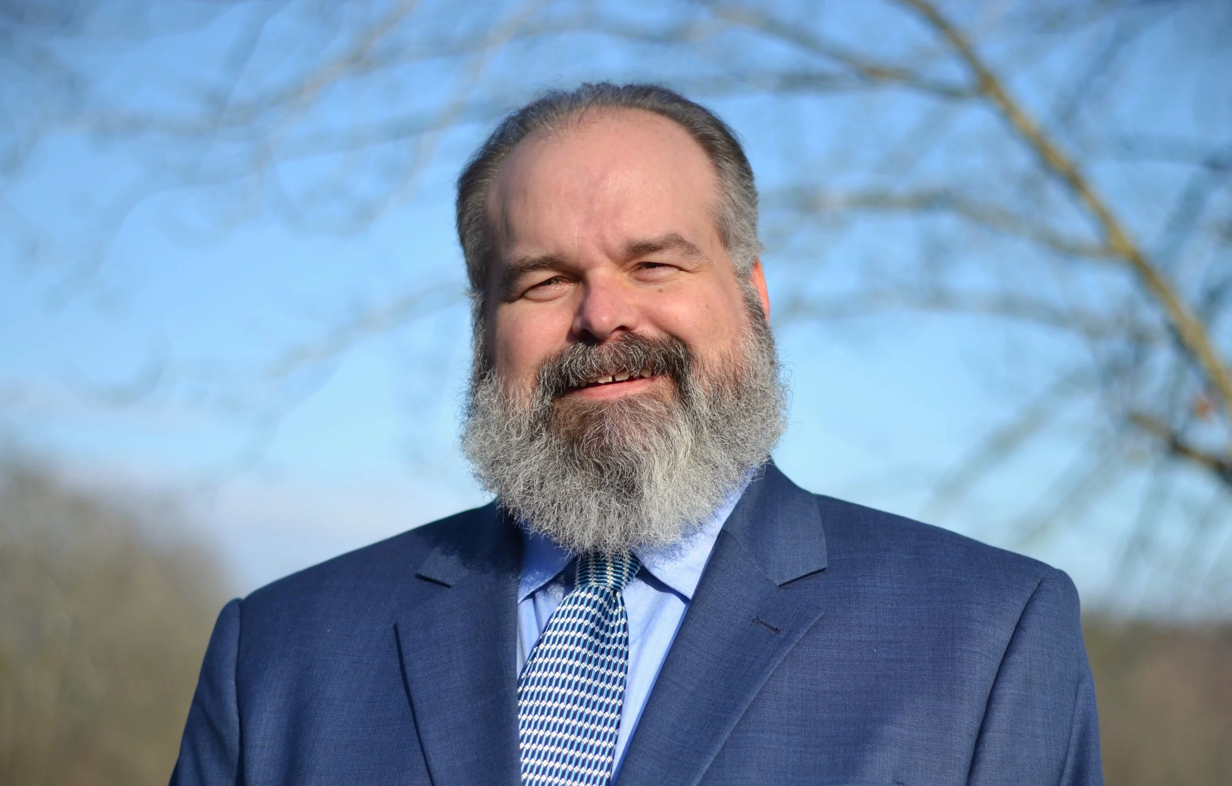 A photo of Stephen Hood, a white man with a beard wearing a blue suit. He's smiling and standing outside with trees behind him.