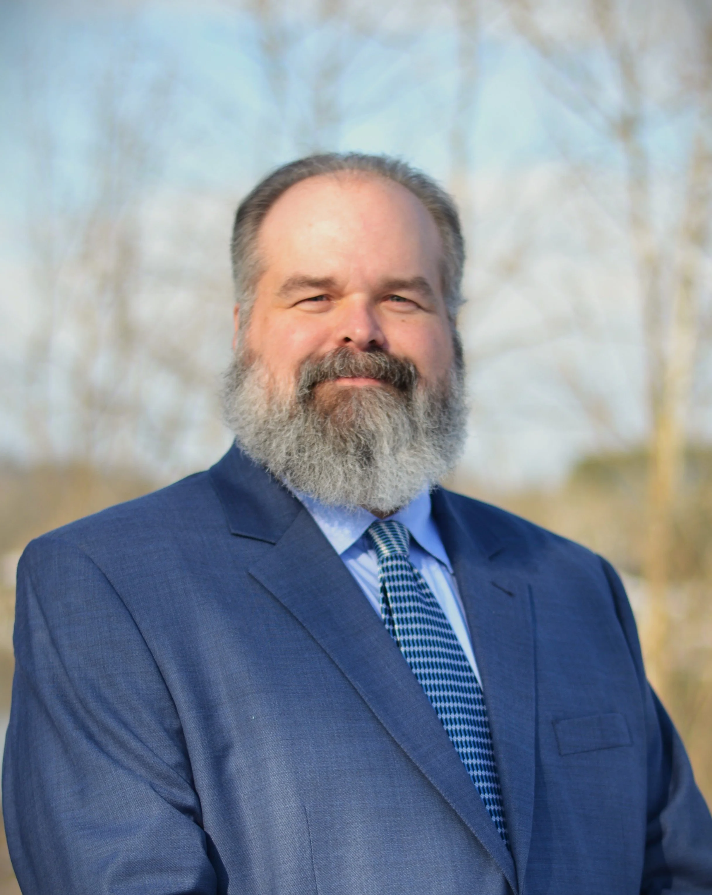 A photo of Stephen Hood, a white man with a beard wearing a blue suit. He's standing outside with trees behind him.
