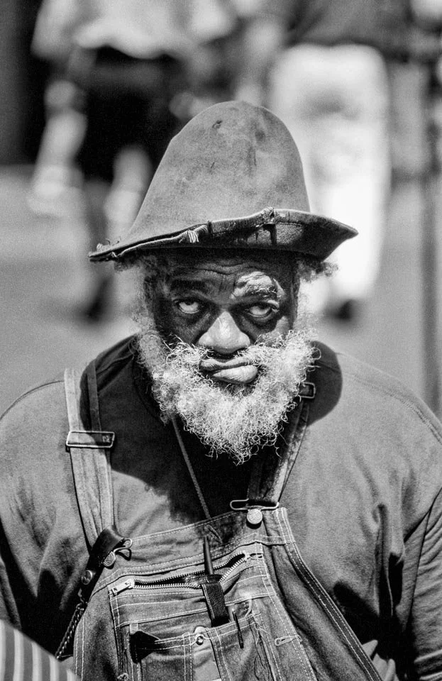 A black and white photo of an elderly man with a beard, wearing a large hat, dark face paint around his eyes, and overalls, looking directly at the camera.