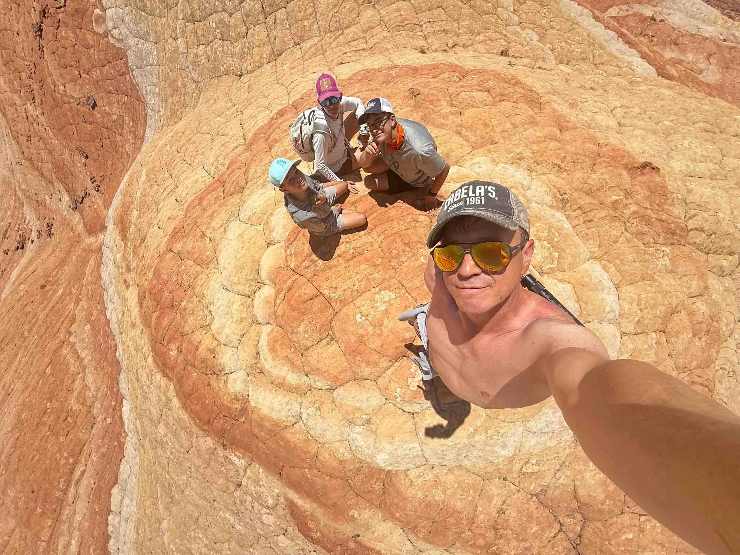 A man taking a selfie with four children sitting on a colorful, layered rock formation in a desert-like environment.