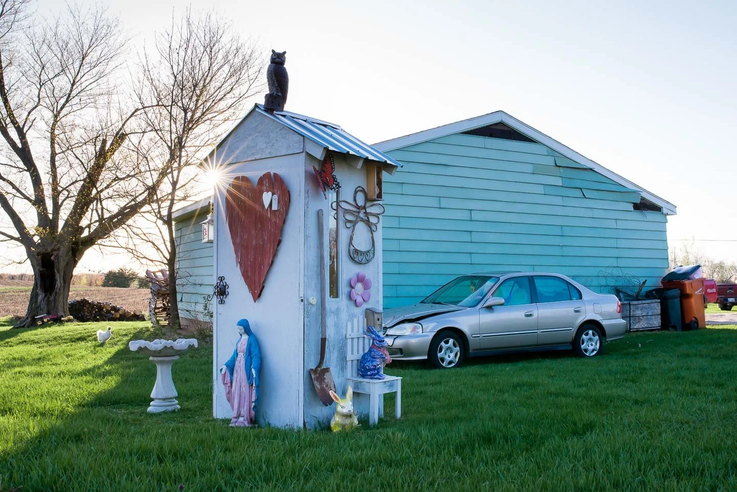 A silver sedan parked on a grassy area in front of a blue house. A small white shed with decorative items, including a large red heart, a blue rabbit, and a statue of the Virgin Mary, is nearby. A tree without leaves is in the background, and the sun