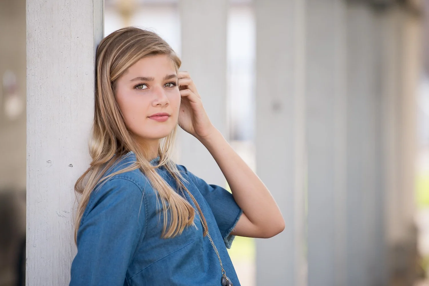A young woman with blonde hair and blue eyes wearing a blue denim shirt and a necklace, standing outdoors next to a white pillar, looking at the camera.