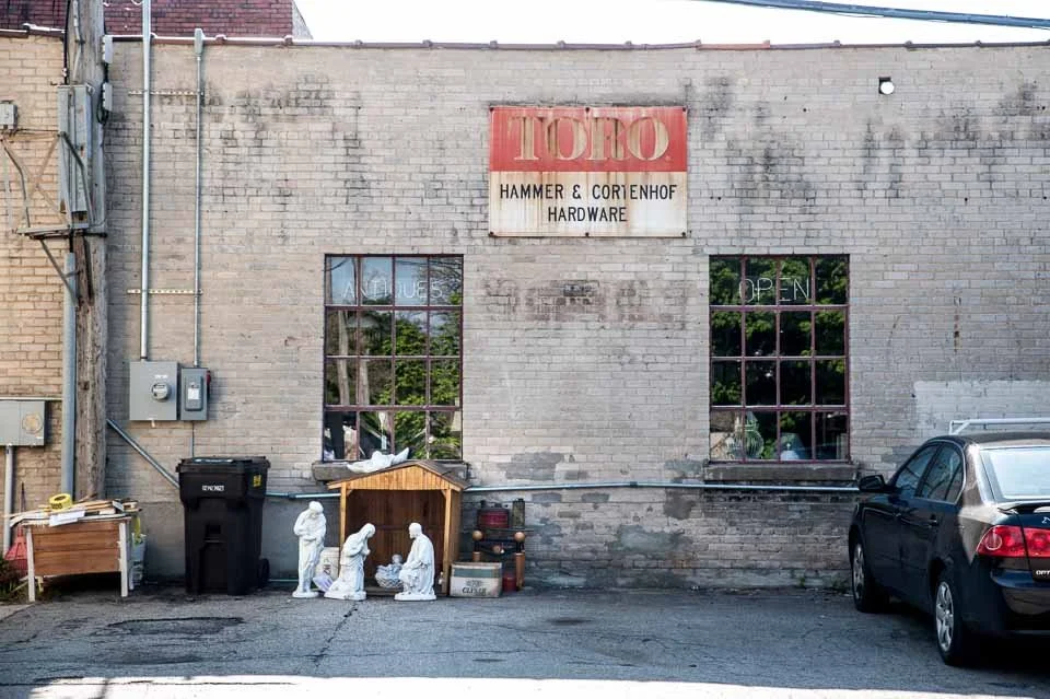 The exterior of a brick building with a red and white sign that reads "TOTO HAMMER & CORTENHOF HARDWARE." There are two windows, a black trash bin, and a small nativity scene with white statues in front. A gray car is parked on the right.