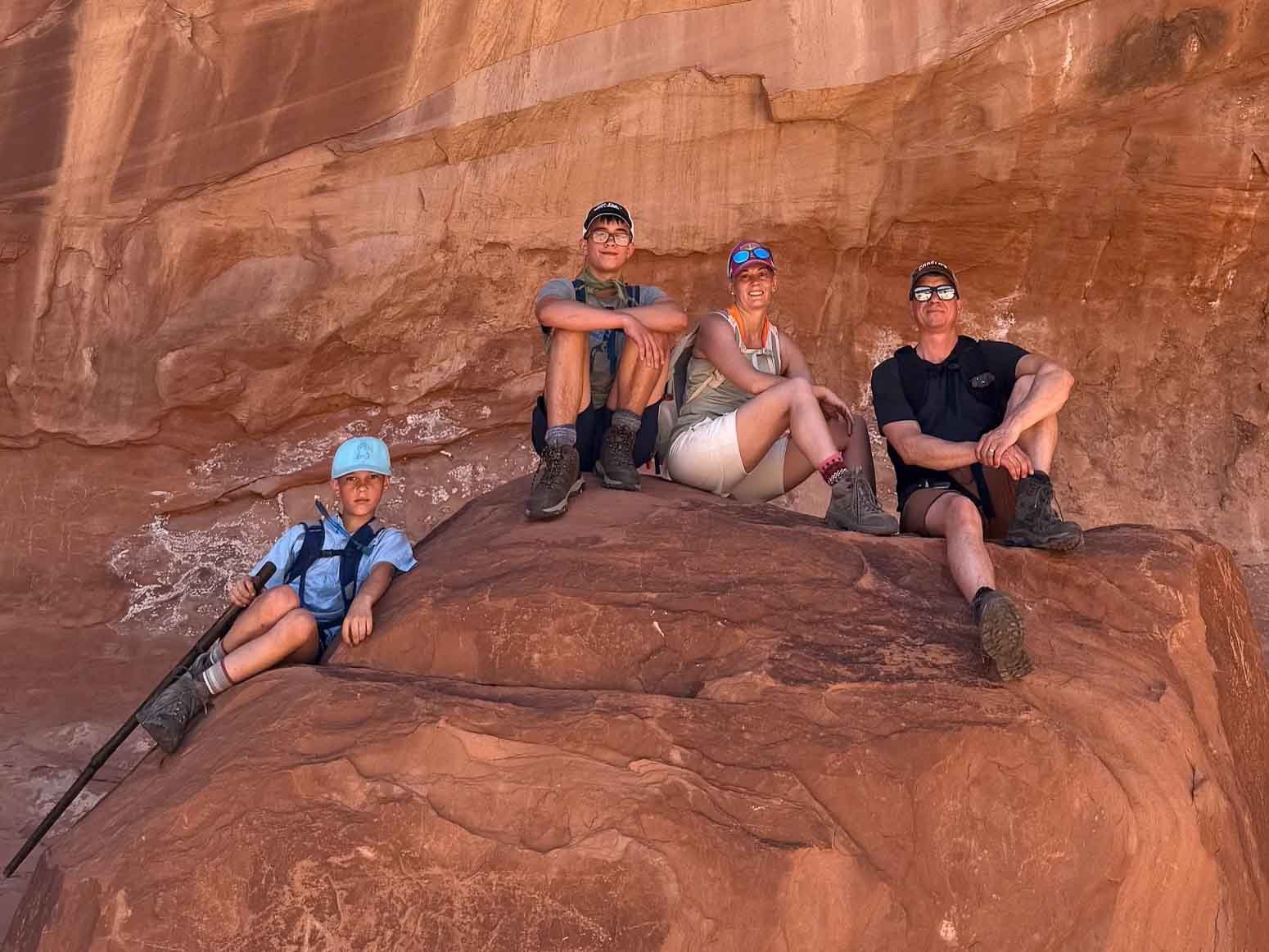 Four hikers sitting and lying on large red rocks in a desert canyon, wearing outdoor gear and sunglasses.