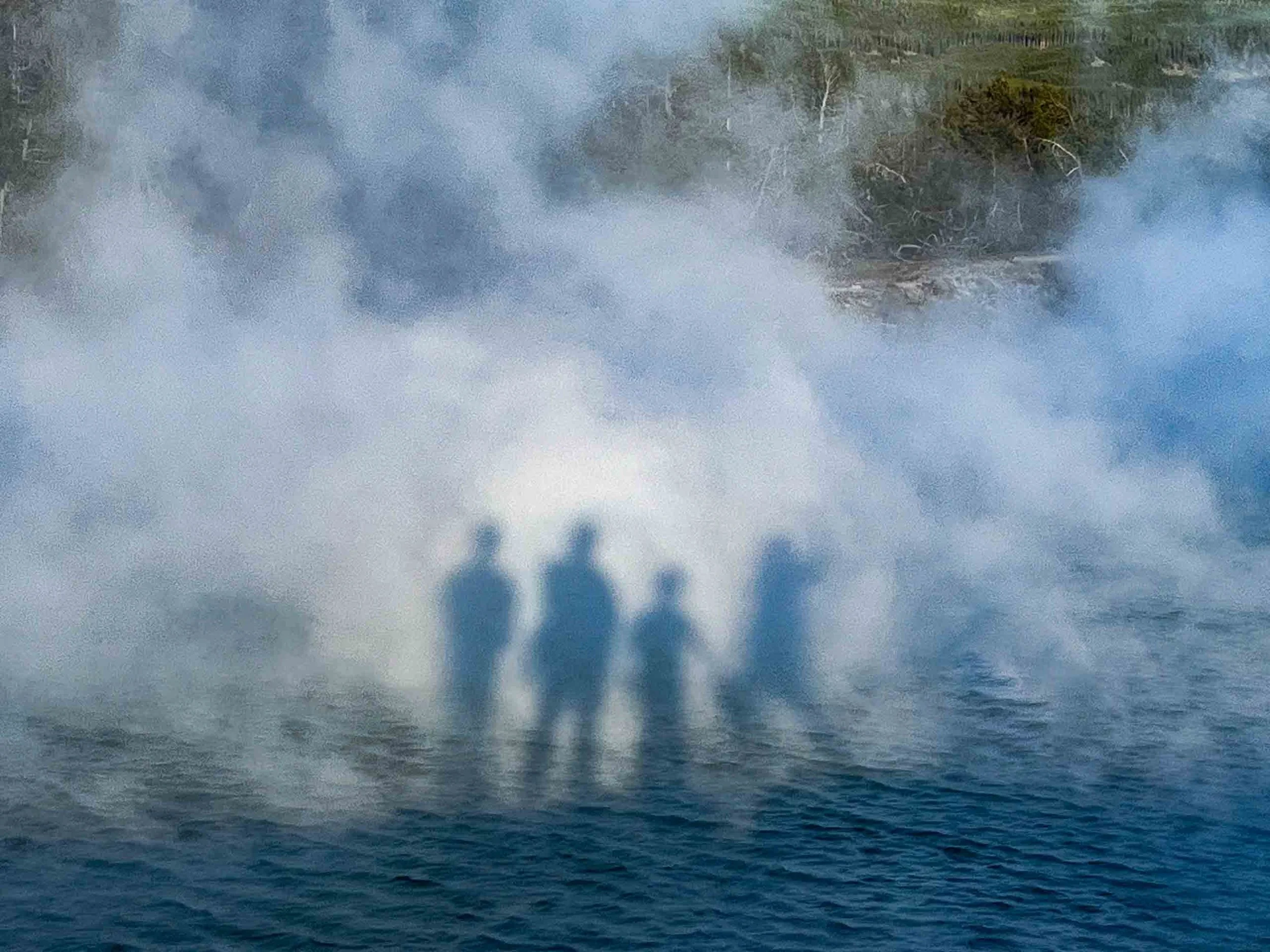Four people explore hot springs surrounded by steam and lush trees.
