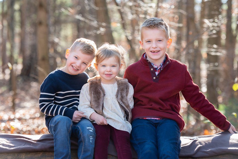 Three children smiling and sitting on a log in a forest with sunlight filtering through the trees.
