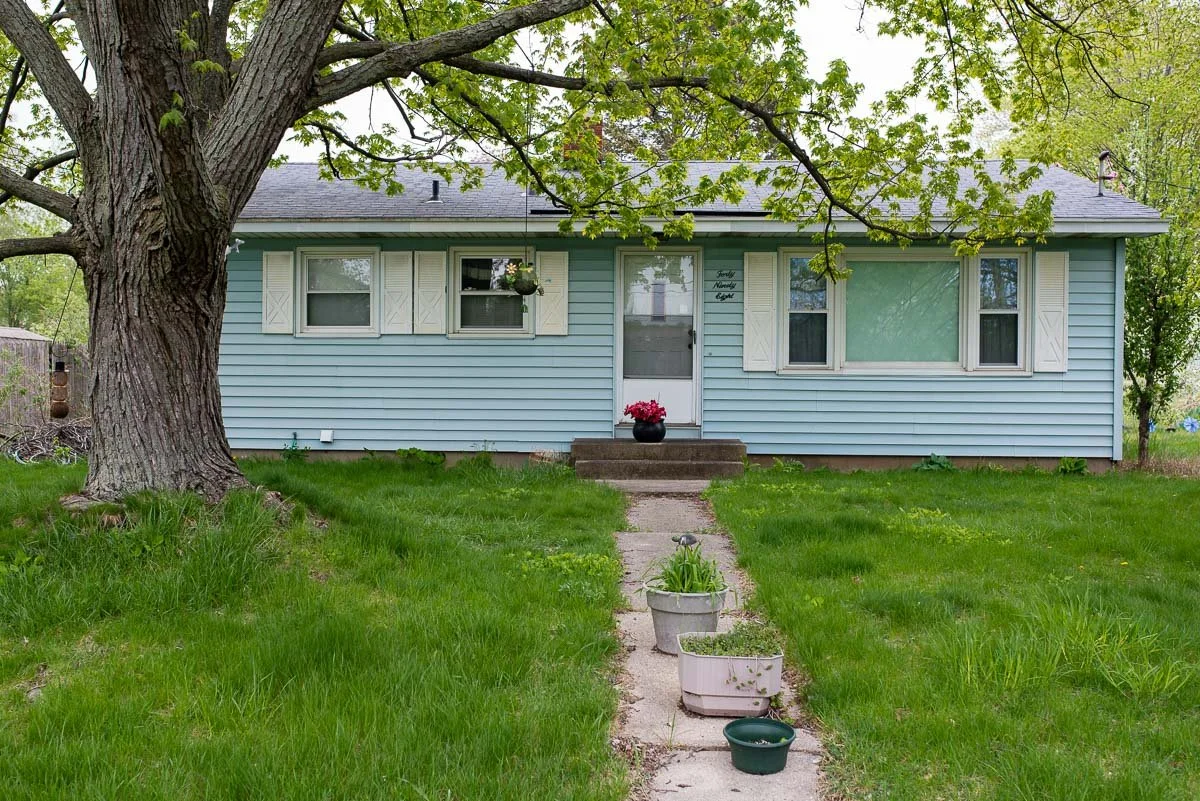 Front view of a light blue house with white shutters, a large tree in the yard, and a concrete walkway with potted plants leading to the front door.