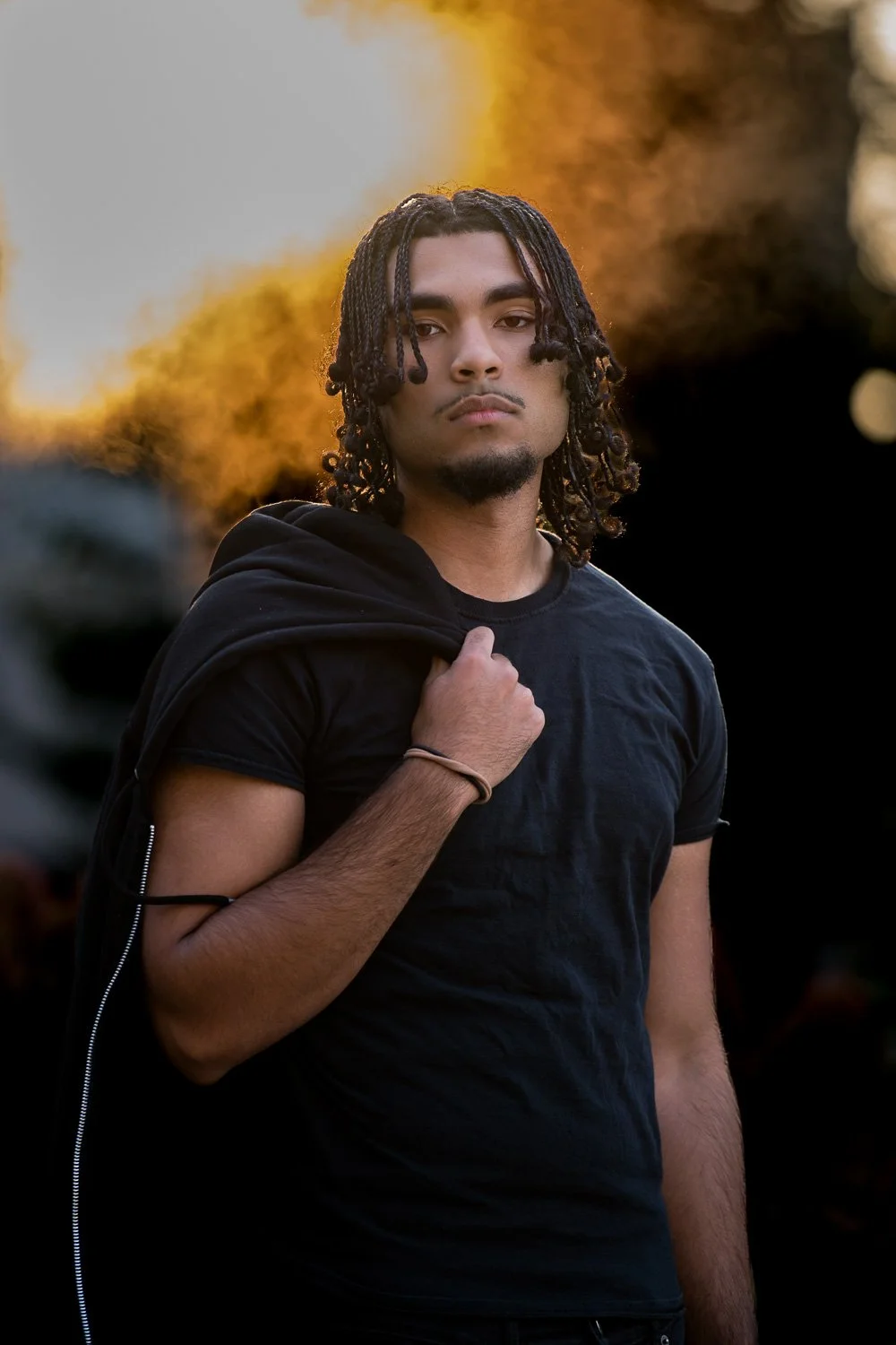 A young man with curly, shoulder-length hair stands outdoors at sunset, wearing a black T-shirt and carrying a black hoodie over his shoulder.
