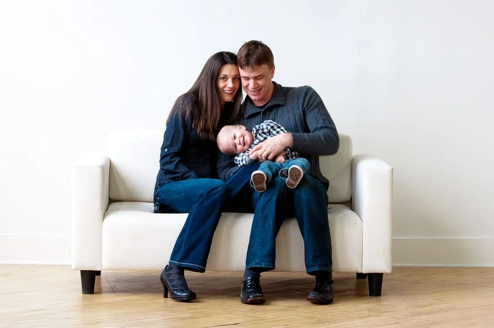 Family of three sitting on a white sofa, smiling and playing with a baby
