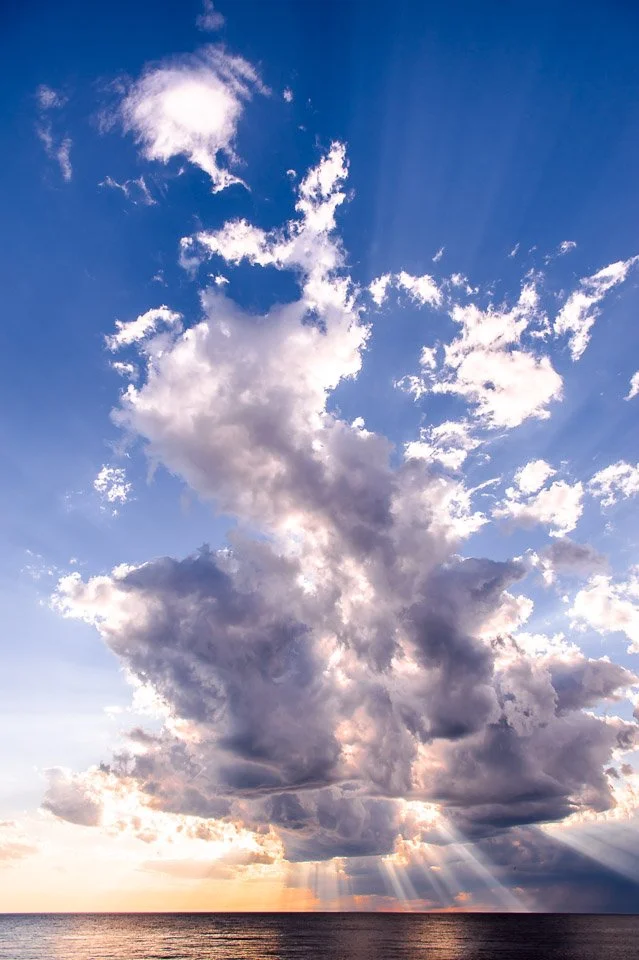 Sky with large, dense clouds and sun rays peeking through at sunset over the ocean.