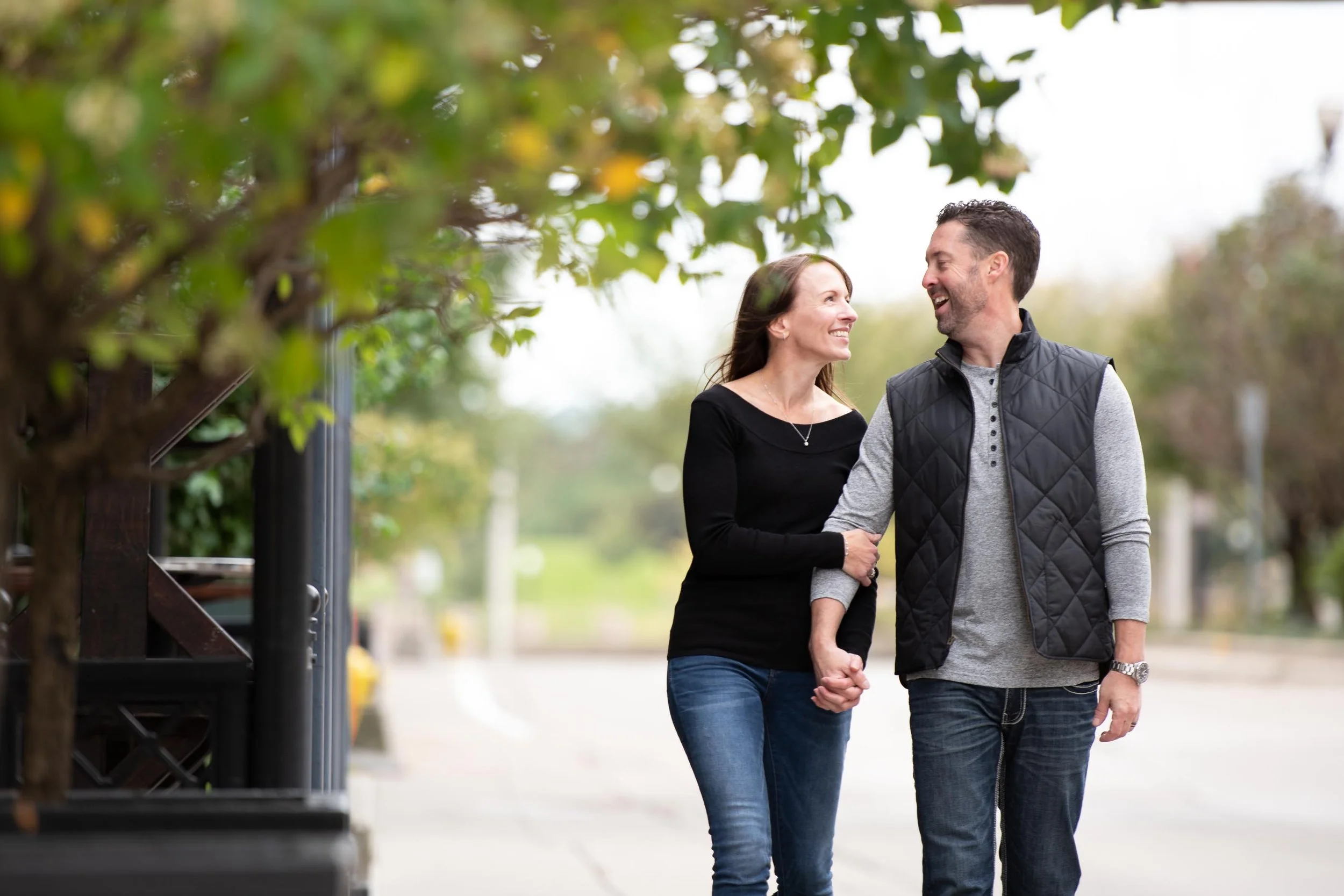 A happy couple walking hand in hand on a city street, smiling and looking at each other, surrounded by trees.