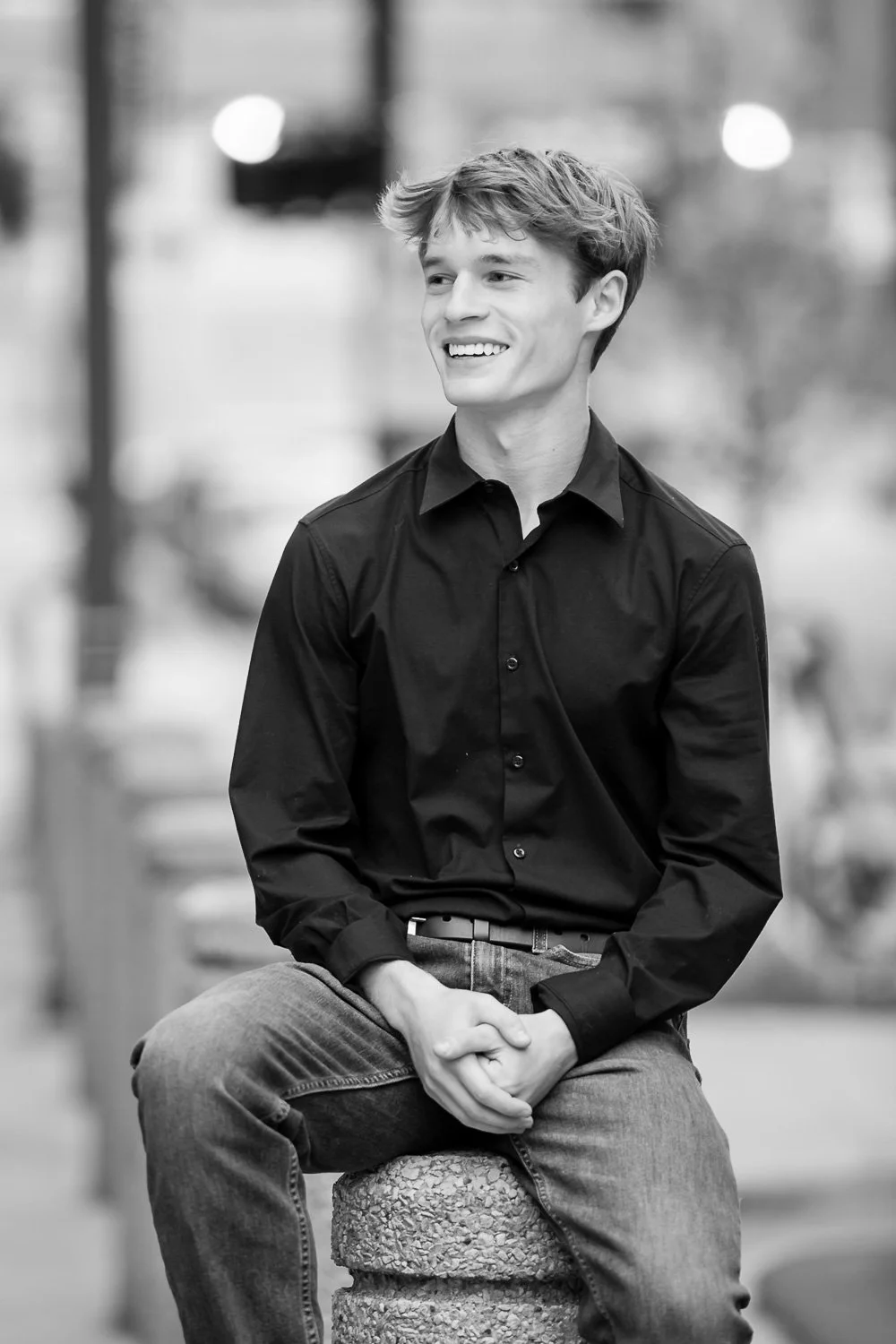 A young man with wavy hair, smiling, wearing a black button-up shirt and jeans, sitting on a textured concrete post in an outdoor urban setting, in black and white.