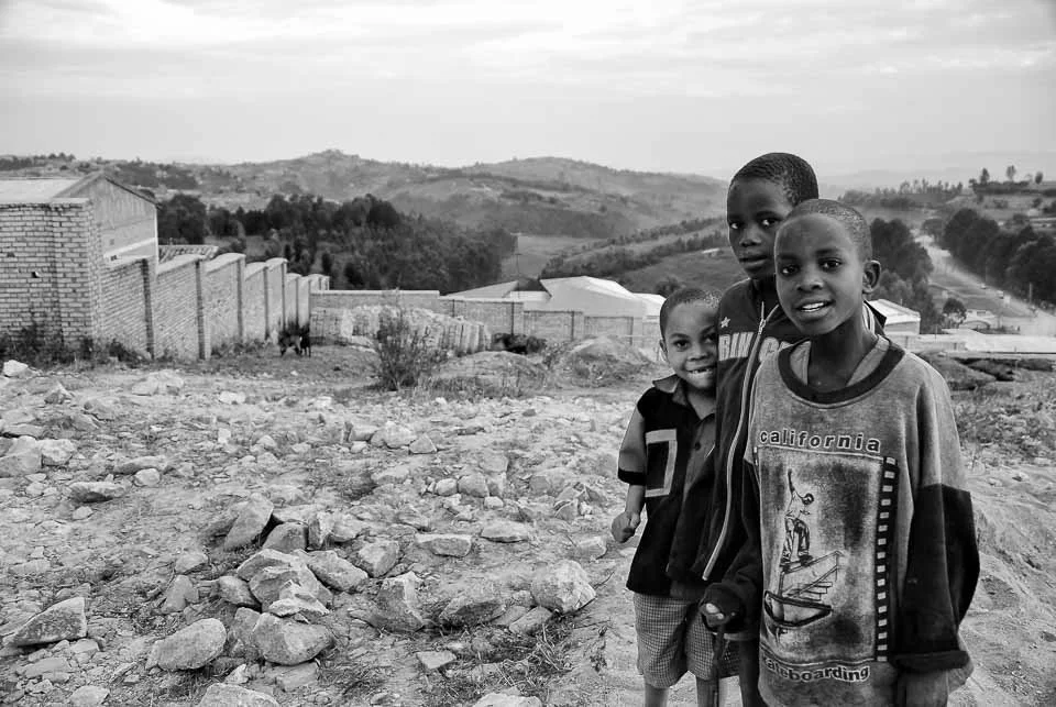 Three young boys standing on rocky dirt ground outdoors with a rural landscape and hills in the background.