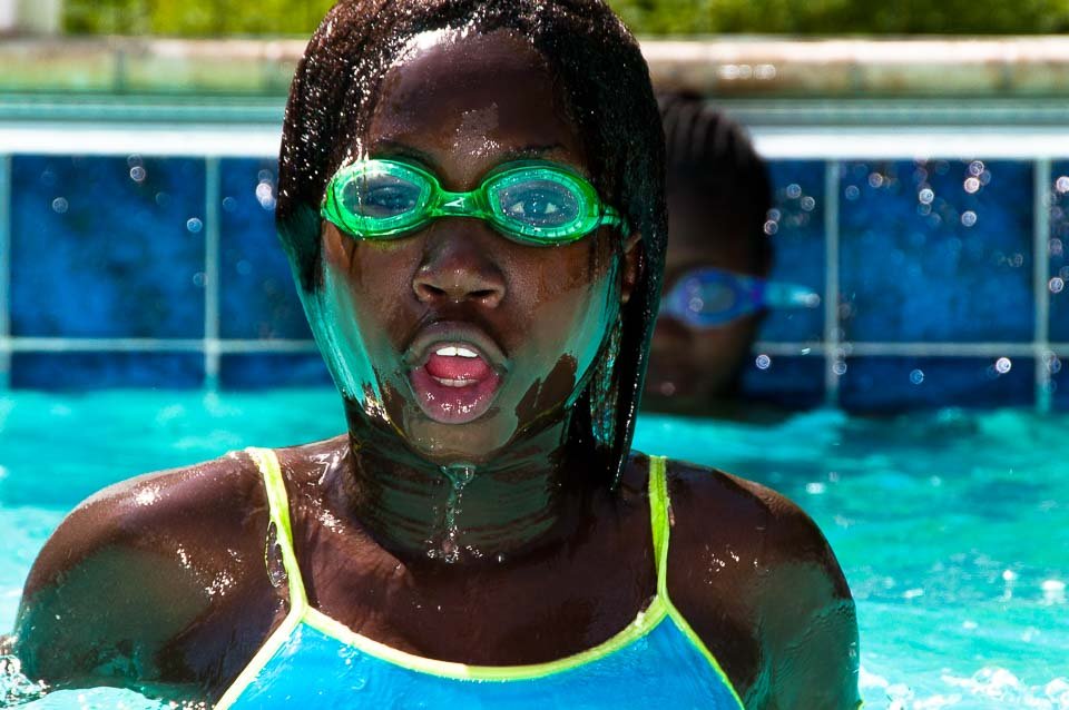 Young girl swimming in a pool wearing green goggles and a blue swimsuit, with another swimmer visible in the background.