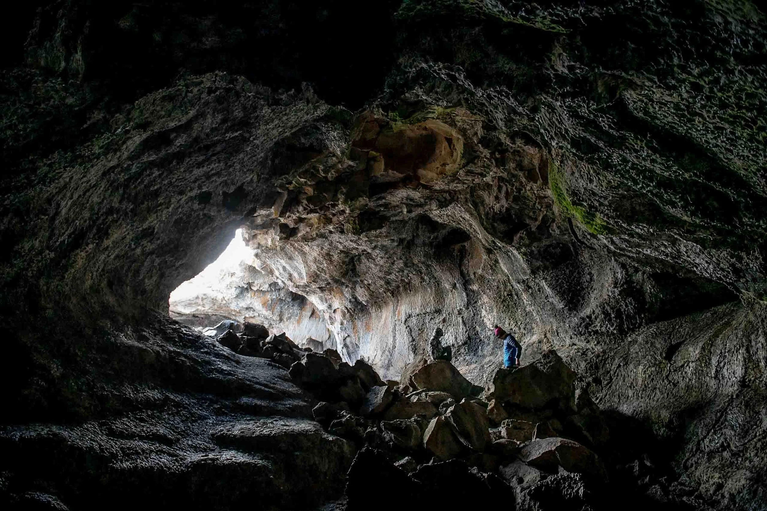 People exploring the inside of a dark cave with rocky surroundings and a bright opening in the background.
