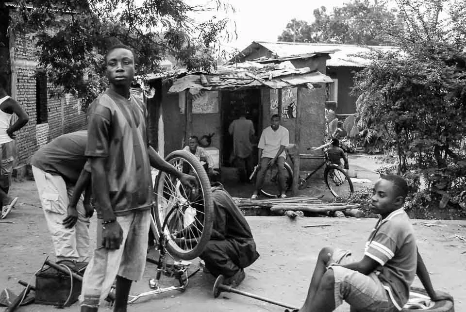 Children outdoors in a neighborhood, with some working on a bicycle and others standing or sitting nearby, in front of a small, dilapidated building.