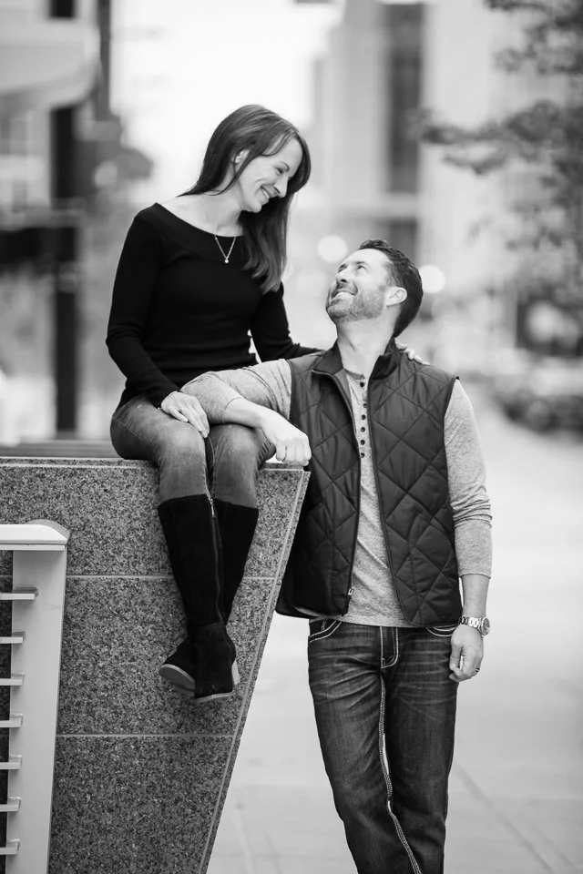 A woman sits on a ledge while smiling at a man standing next to her, looking up at her, in an urban setting.